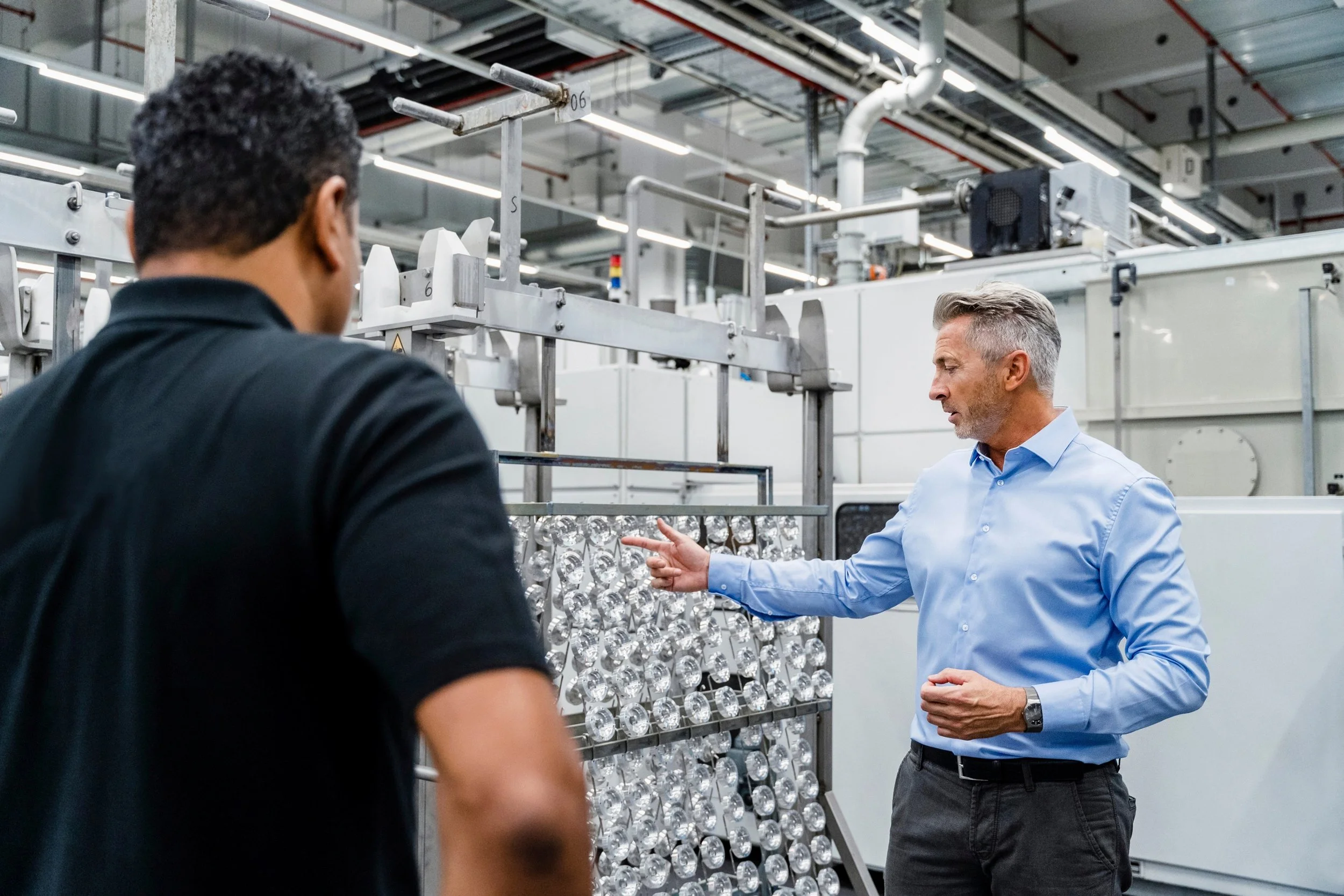 Two men are in a factory, with one of them explaining something. The man on the right has gray hair and is wearing a light blue shirt, while the man on the left has dark curly hair and is wearing a black shirt. The factory environment has machinery and a rack of clear glass or plastic objects.