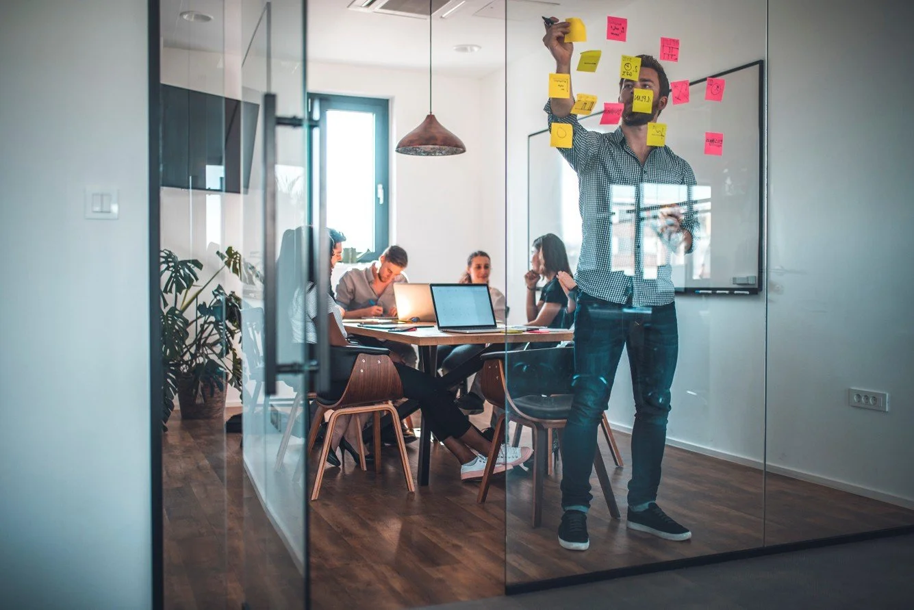 A team of young professionals in a meeting room with one team member adding sticky notes to a window