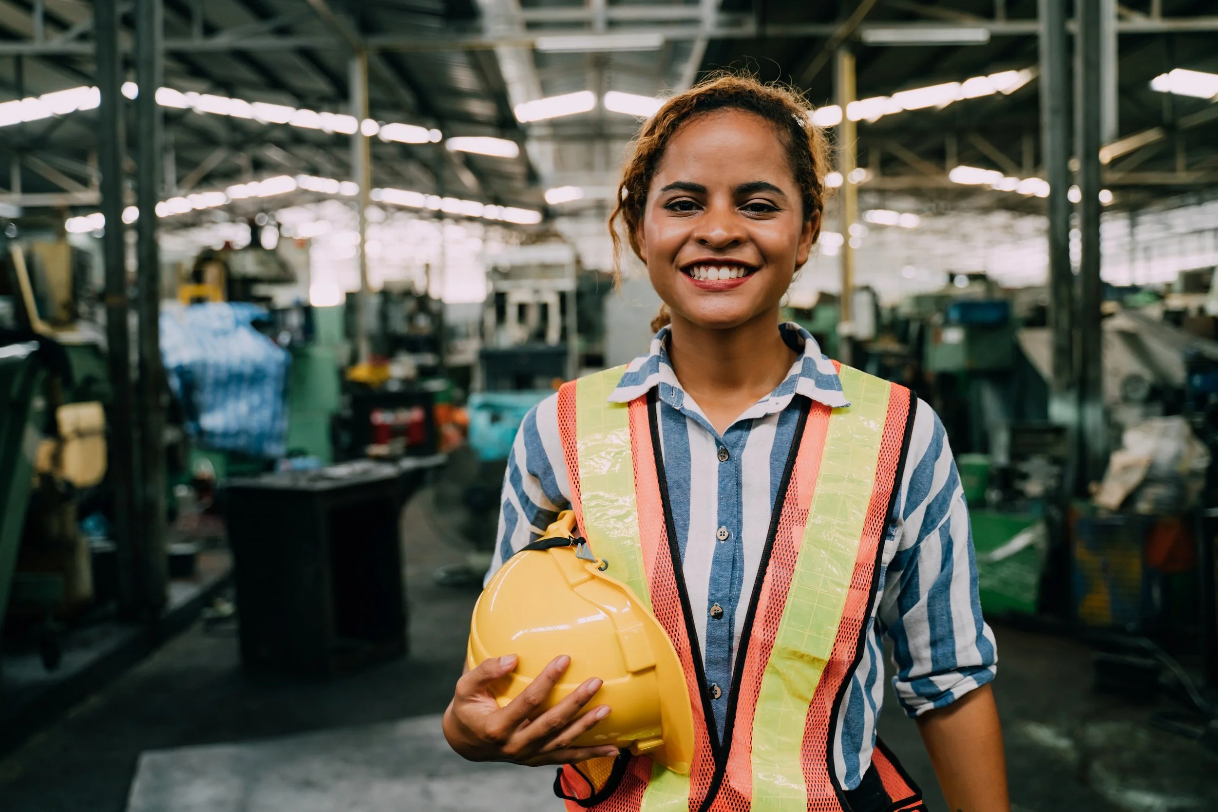 A woman in a striped shirt and safety vest holding a yellow safety helmet in an industrial warehouse.