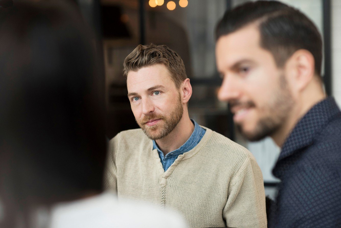 Two men engaged in a conversation, one with blonde hair and a beard wearing a beige sweater, the other with dark hair and a beard wearing a dark patterned shirt, in a cozy indoor setting.