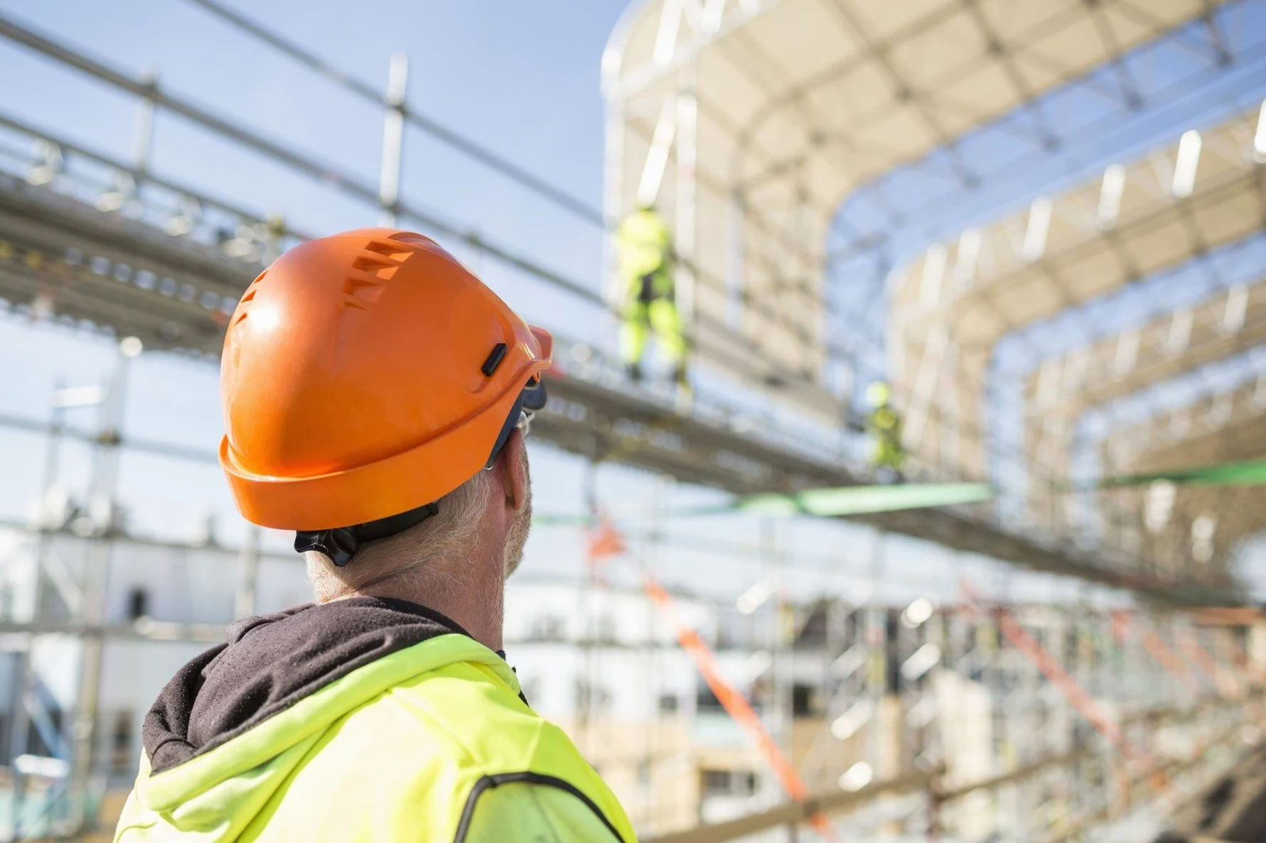 A construction worker in a hard hat and hi-vis clothing, looking up at other workers on scaffolding