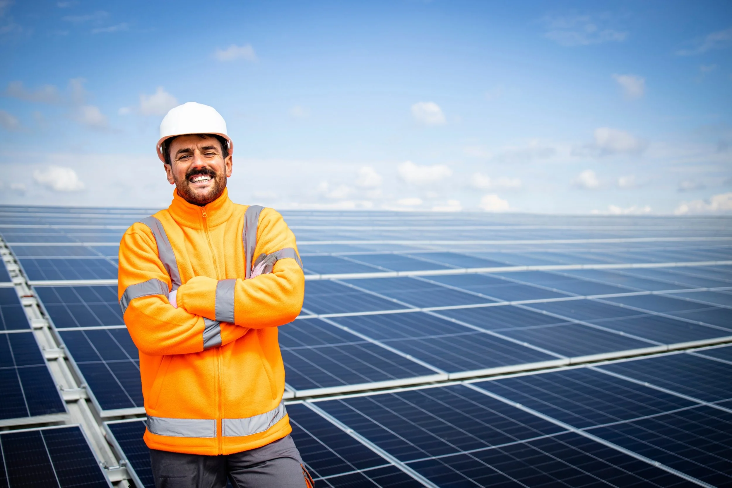 An industrial energy worker, wearing a hard hat and a hi-vis jumper, standing in front of a field of solar panels