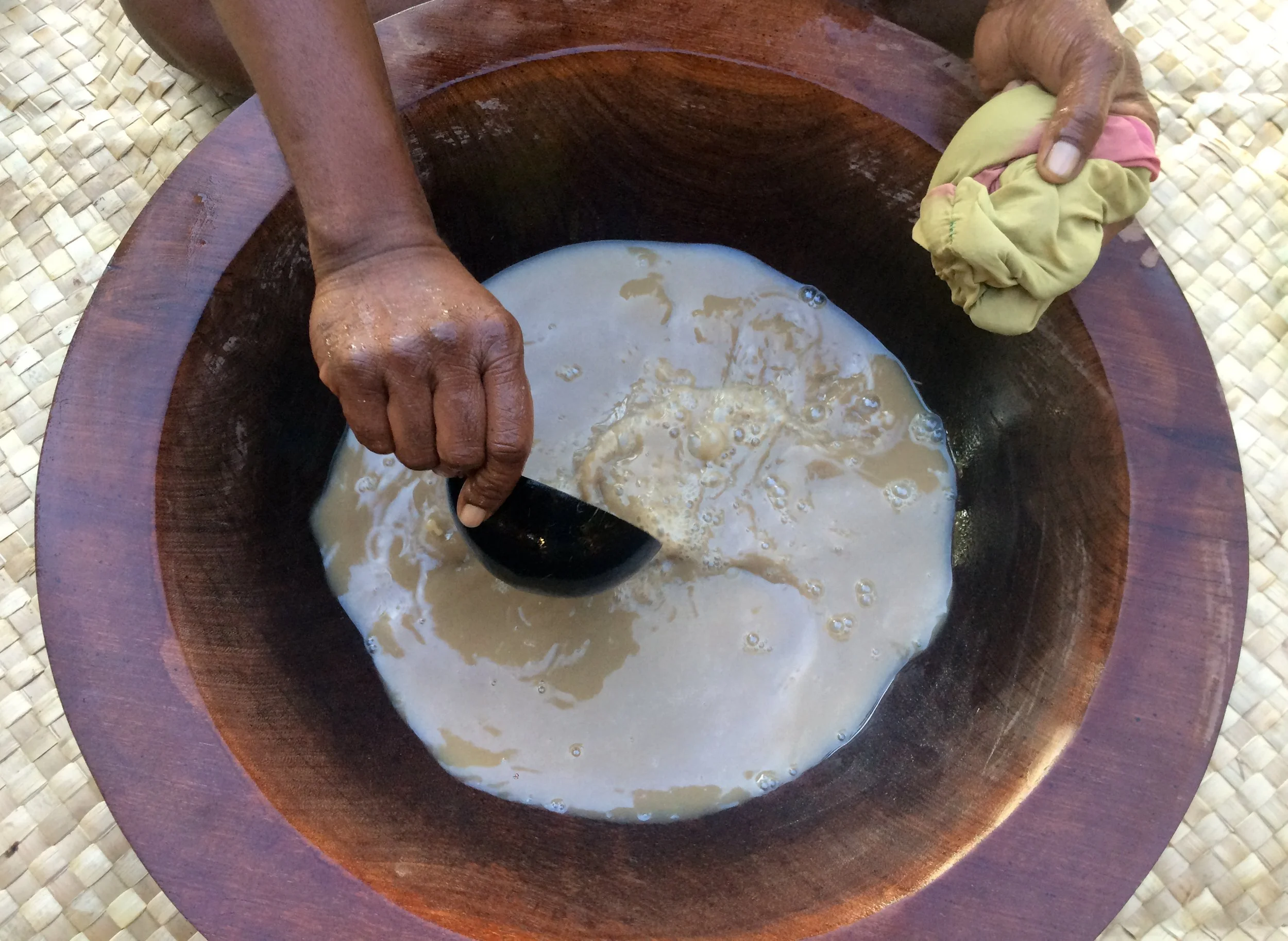 Hands of an elderly person holding a cloth and a ladle over a wooden bowl with liquid