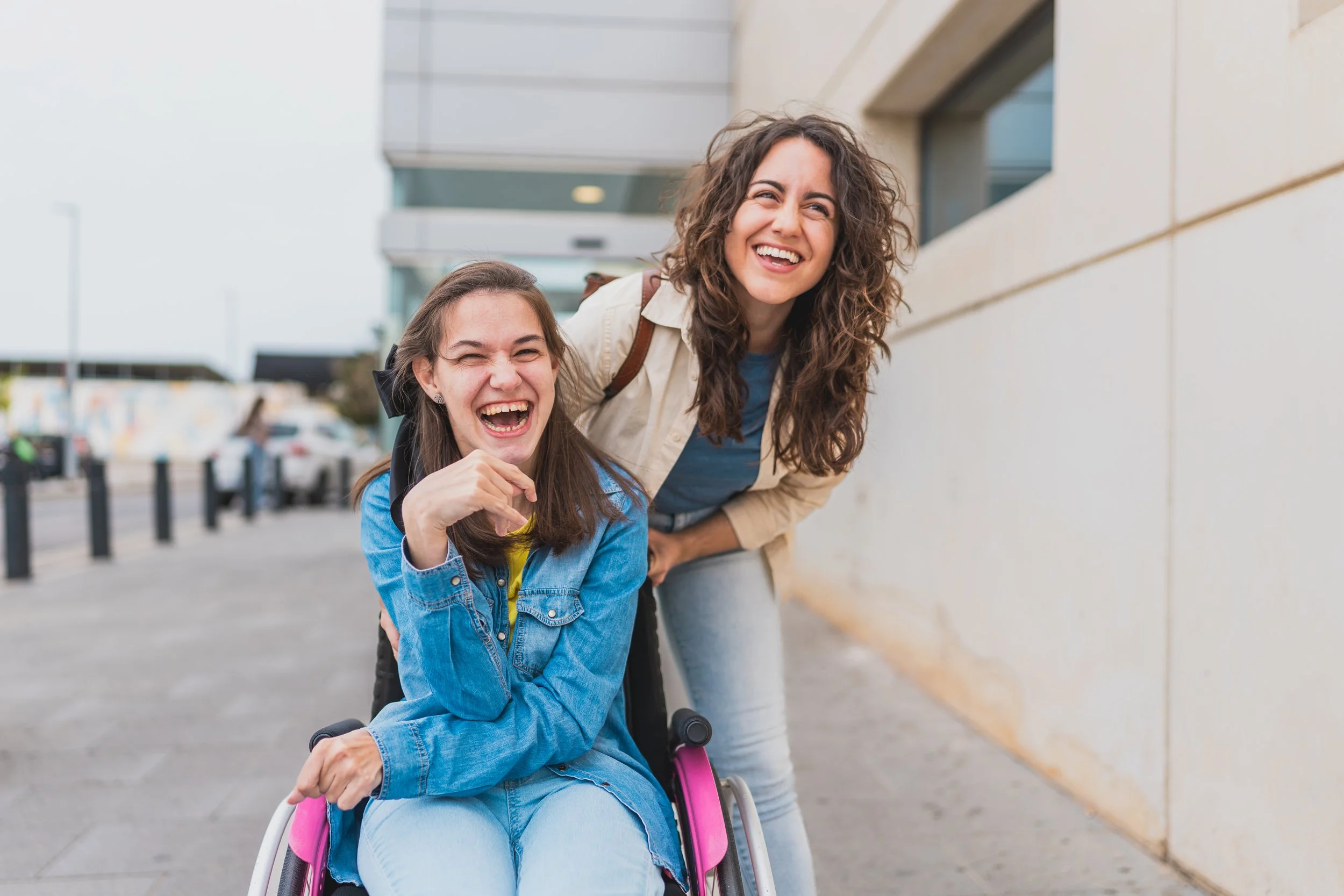Two women laughing and enjoying together outside, one standing and leaning over a wheelchair where the other woman is sitting, with a modern building and parking lot in the background.