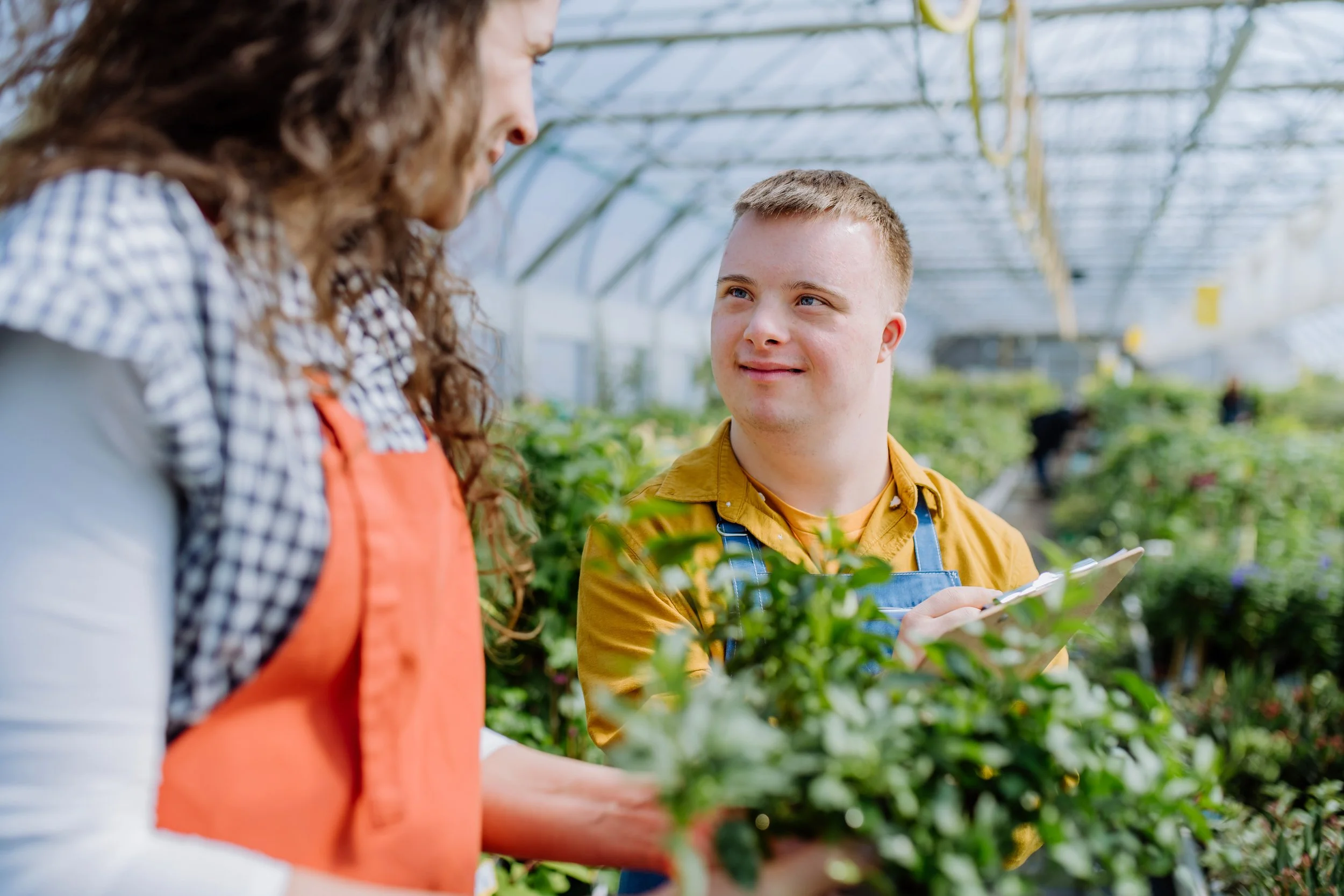 A young greenhouse worker with a clipboard, standing amongst plants, looking at his colleague