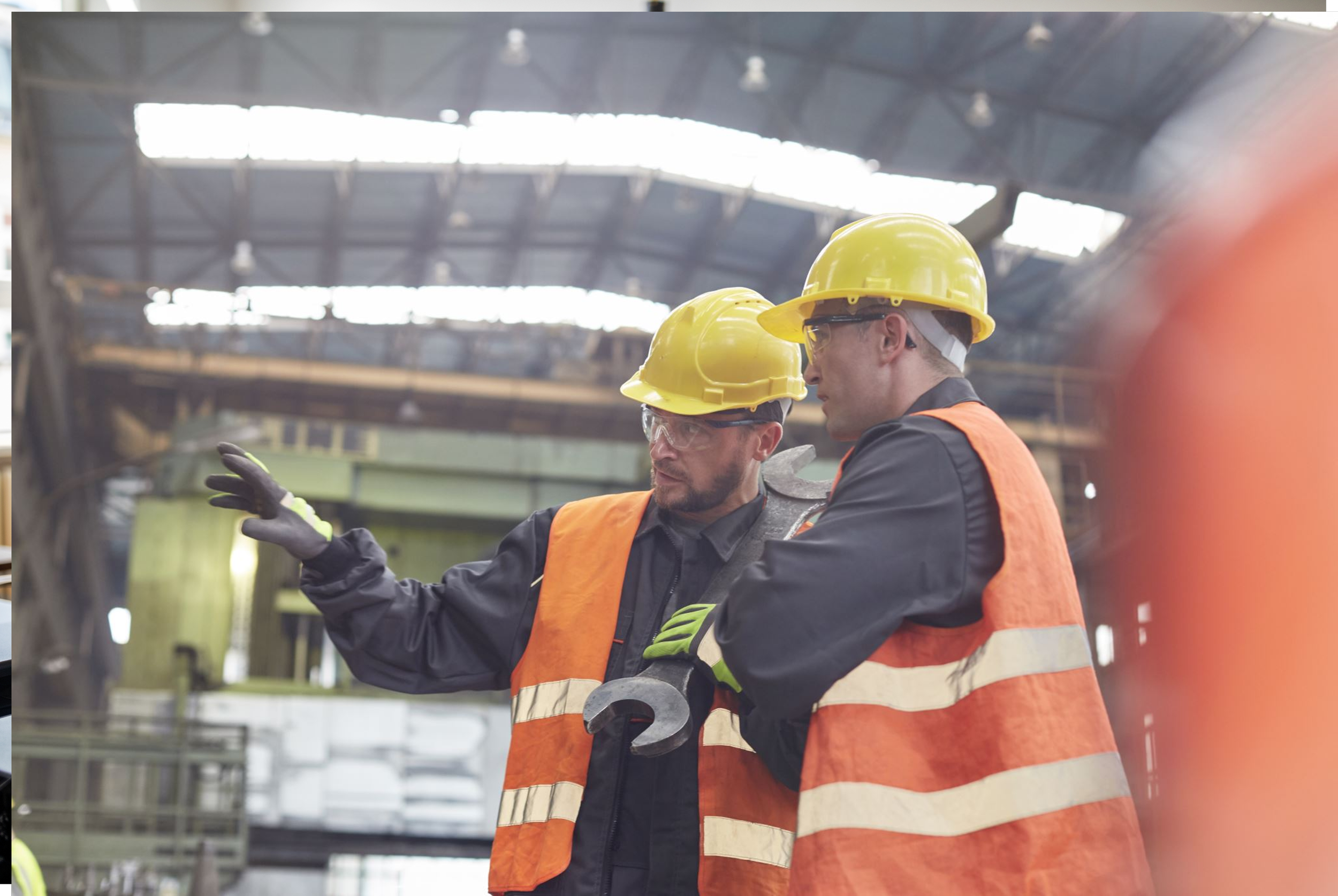 Two industrial workers, wearing hard hats and hi-vis vests, in conversation in an industrial setting