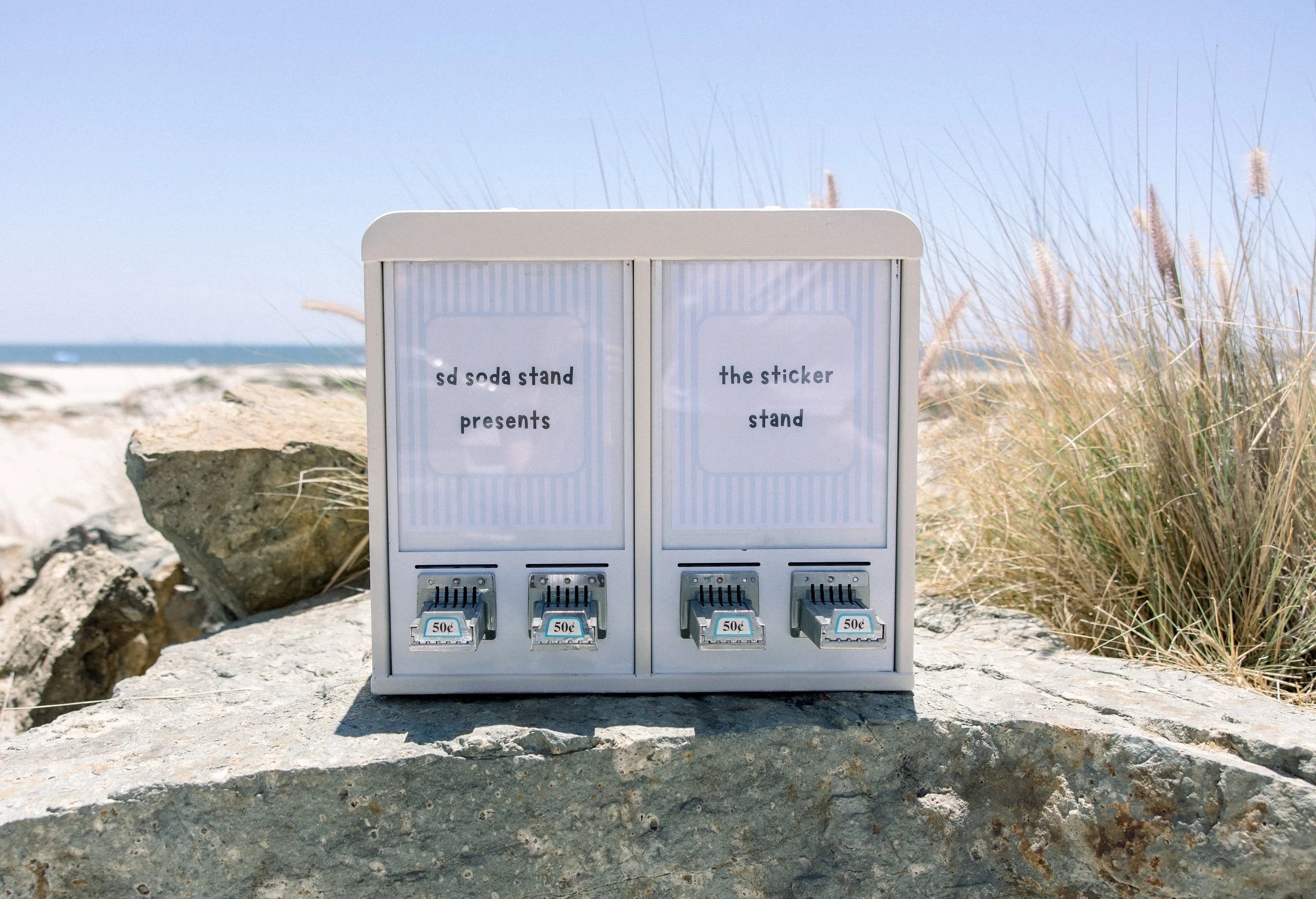 A small vending machine labeled 'soda stand presents' and 'the sticker stand' with four coin slots, located outdoors on a rocky surface near beach grass and rocks, with the ocean and sky in the background.