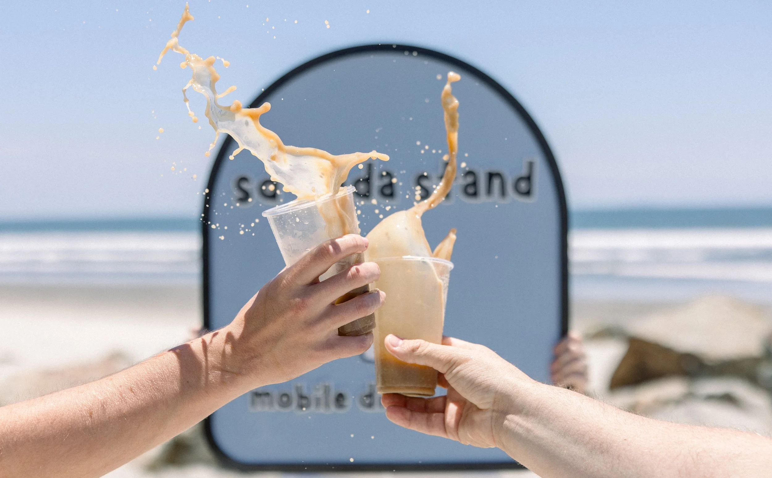 Multiple hands holding ice coffee cups with coffee spilling out, in front of a beach backdrop with a blue sign that reads 'sand's mobile'.