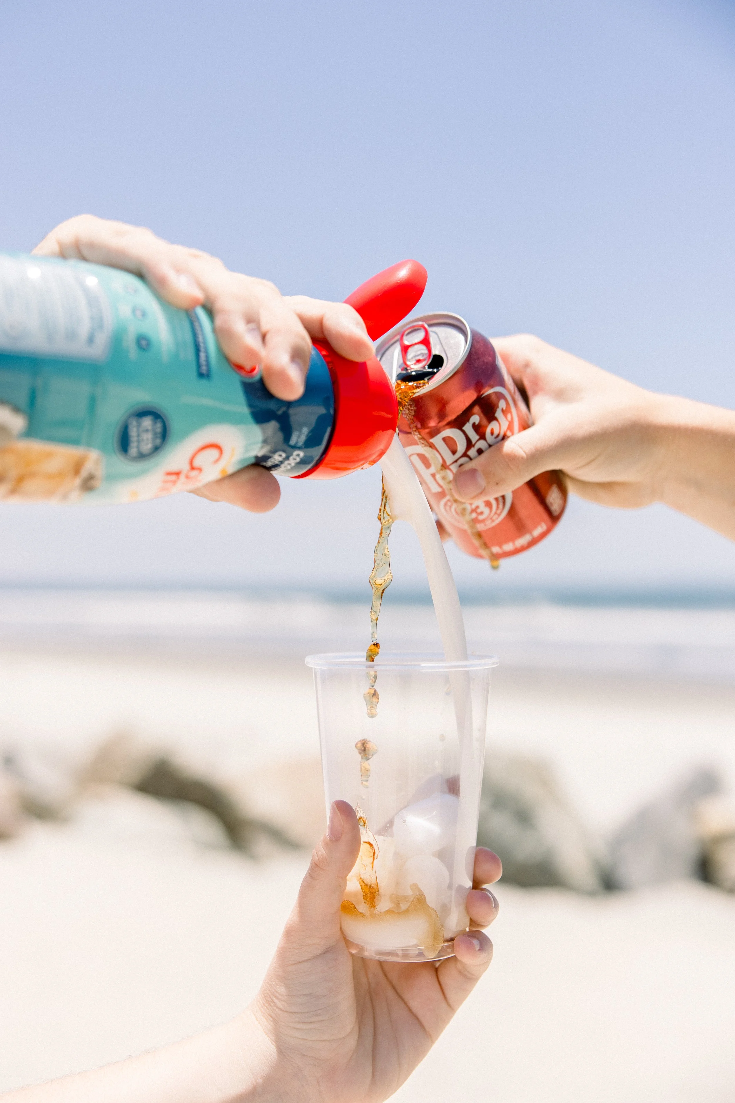Someone holding a clear plastic cup filled with ice and soda while pouring more soda from a can and a bottle, with a beach in the background.
