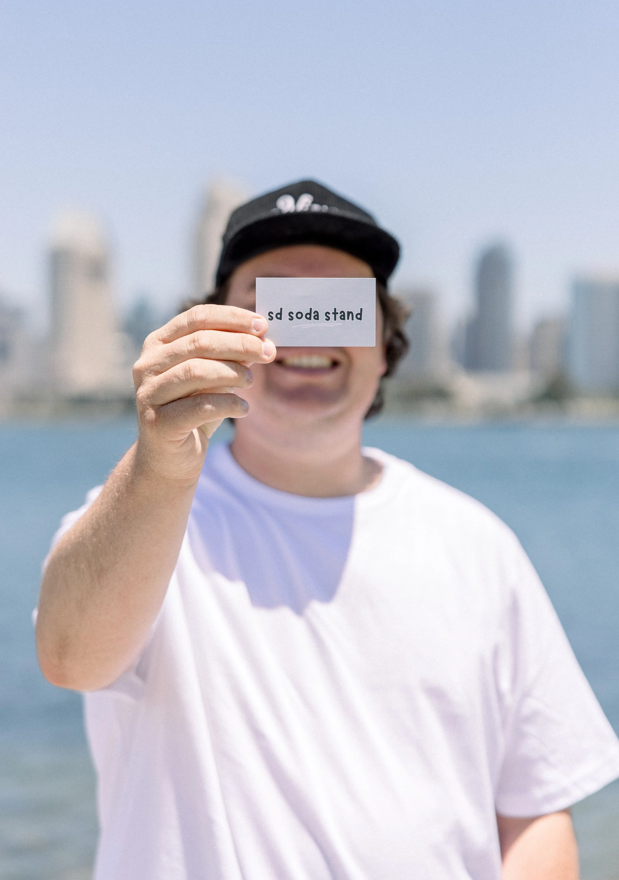 Person holding a sign that says 'sd soda stand' at the beach with skyline in the background.