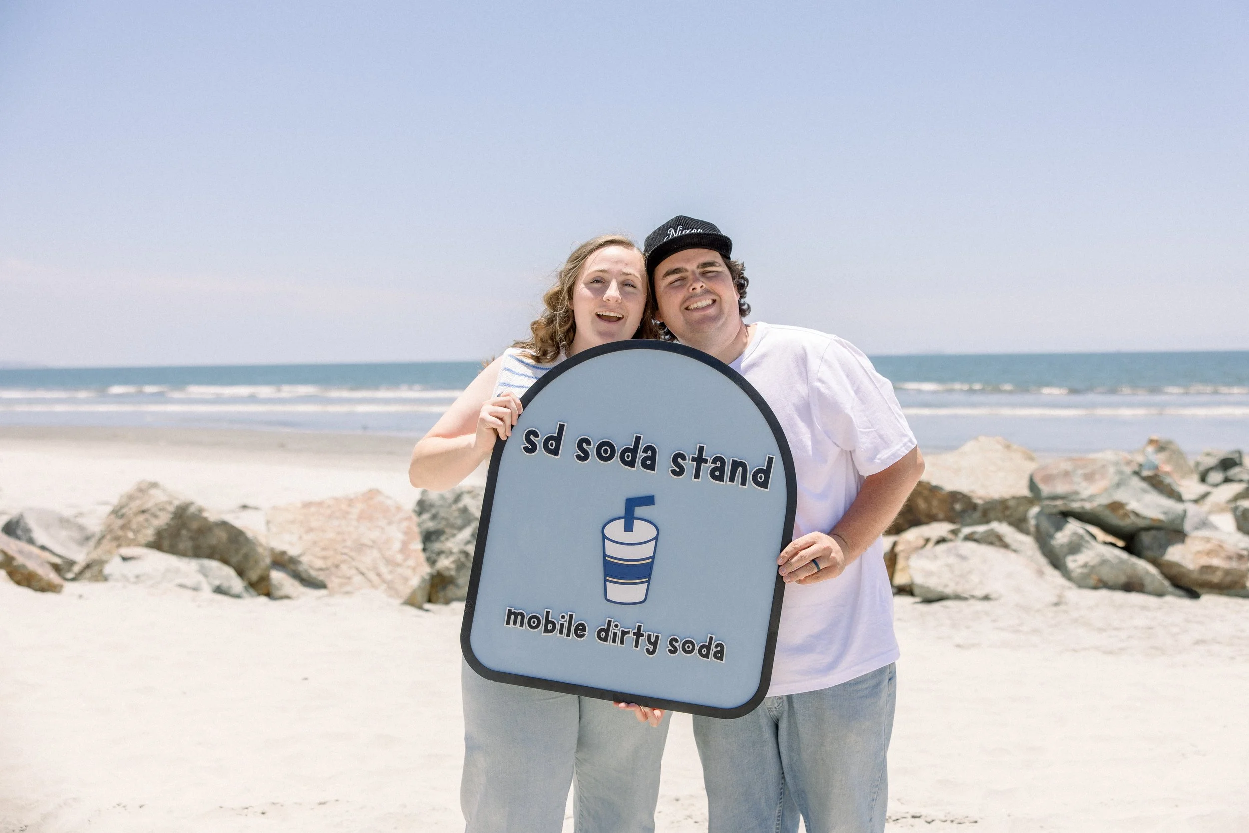 Two people at the beach holding a sign that reads 'sd soda stand mobile dirty soda' with a picture of a soda cup, smiling and squinting in the sunlight.