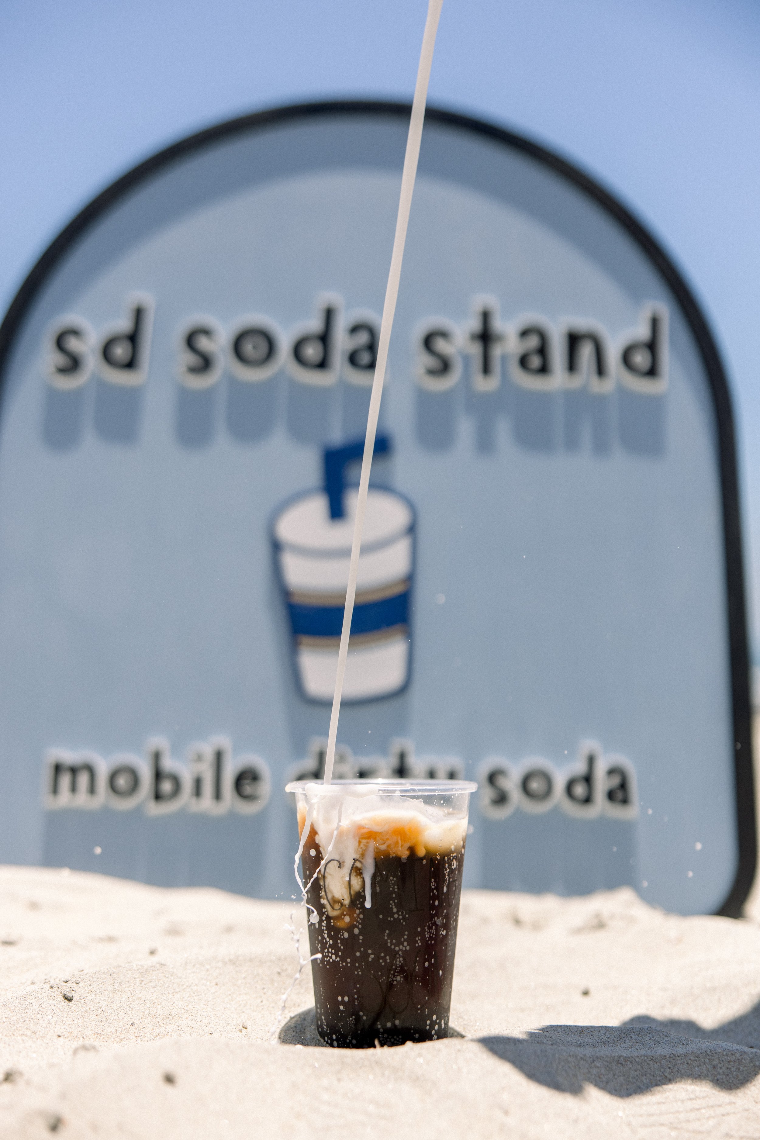 A plastic cup filled with a dark soda and ice cubes, with spilled soda on the outside, placed on sandy ground with an outdoor background.
