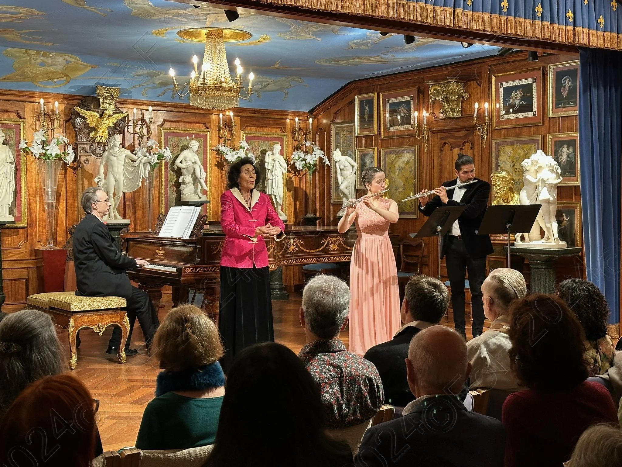 A classical music performance in an elegant, wood-paneled room. Four musicians, including a pianist, two flutists, and a singer, perform in front of an audience. The room is decorated with statues, paintings, and a chandelier.