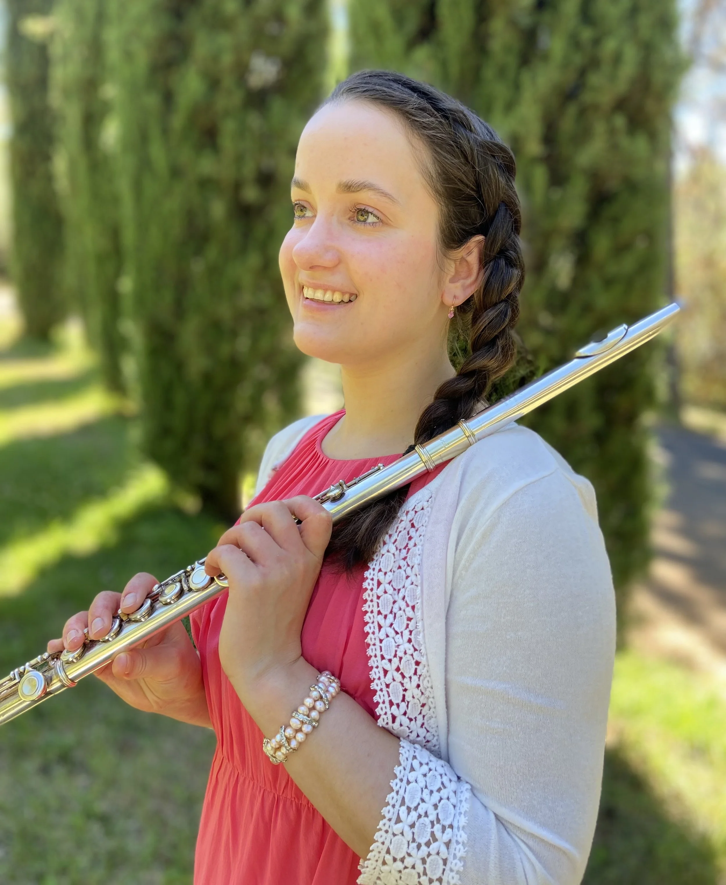 A young woman playing a silver flute outdoors surrounded by greenery, wearing a pink dress, a white cardigan, and pearl jewelry.