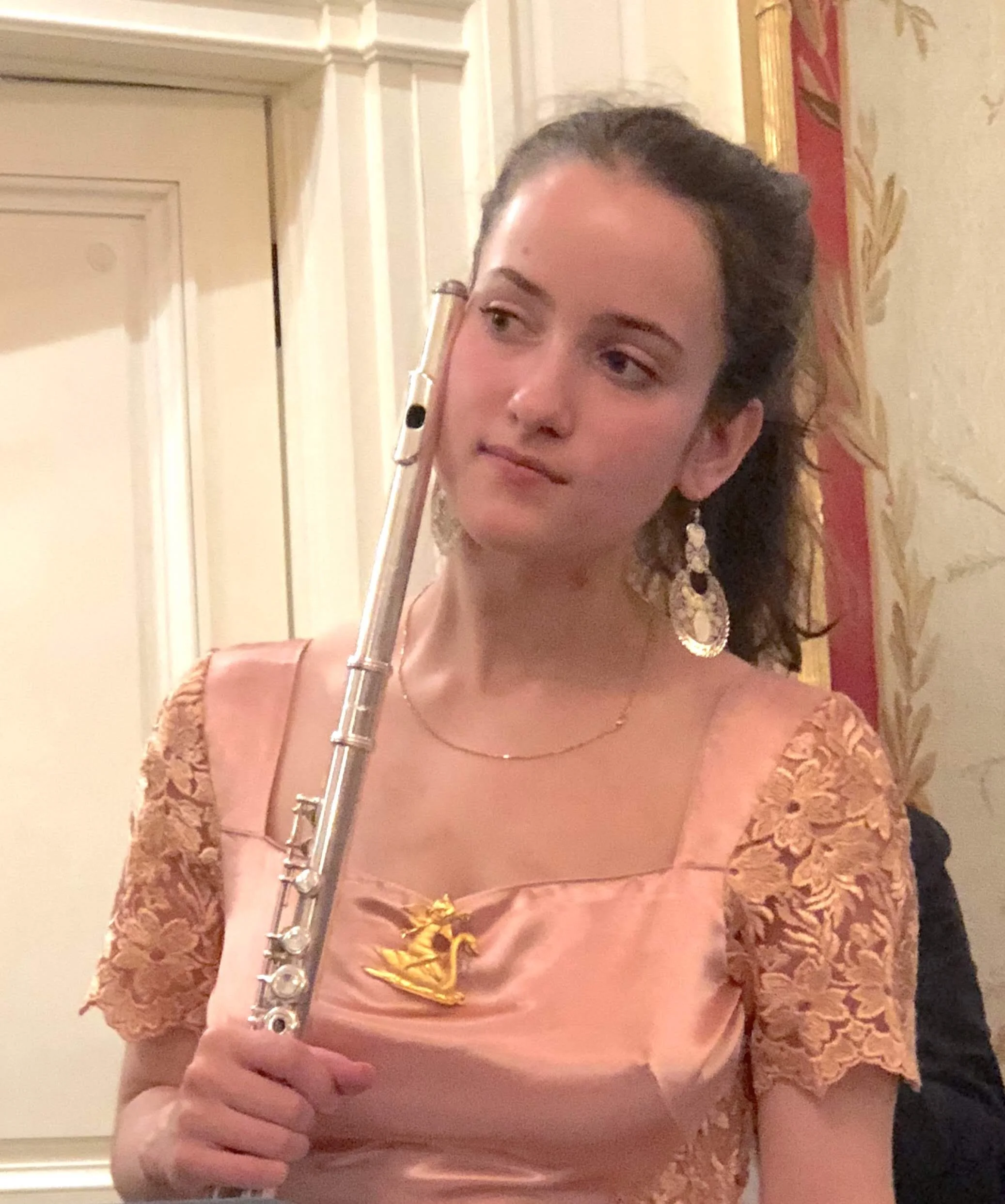 A woman holding a flute, dressed in an elegant dress with lace sleeves, standing indoors with patterned wallpaper and ornate moldings in the background.