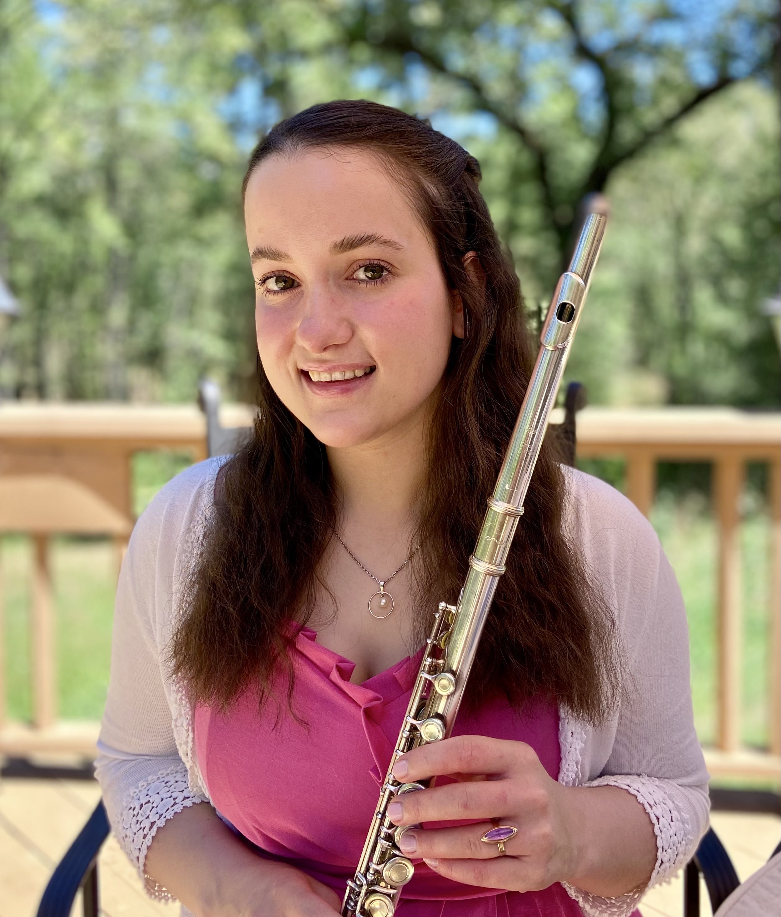 A woman with long brown hair holding a flute outdoors, wearing a pink top and a light-colored cardigan, with a blurred green and blue background of trees and sky.