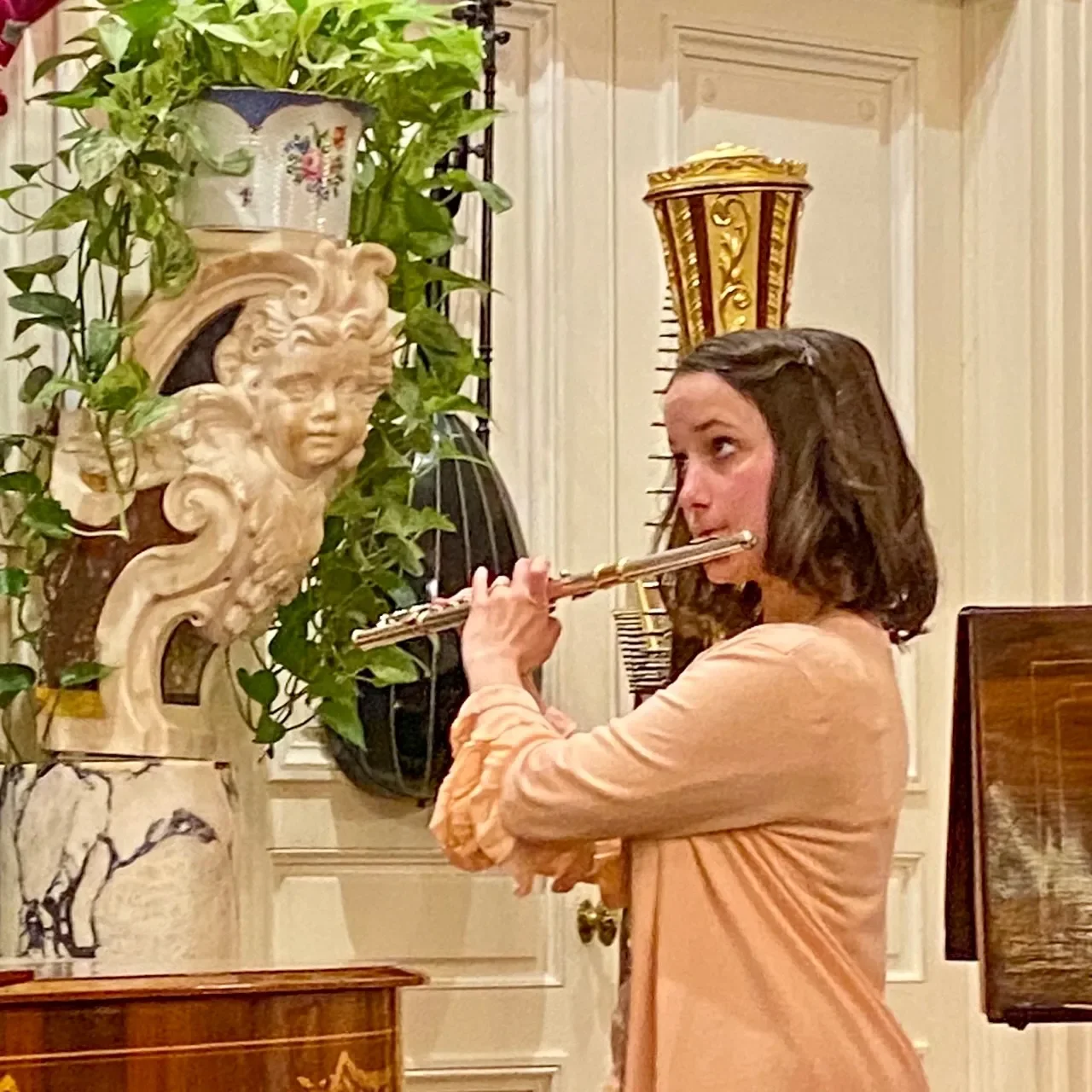 A woman plays the flute in a room decorated with ornate artworks and furniture.