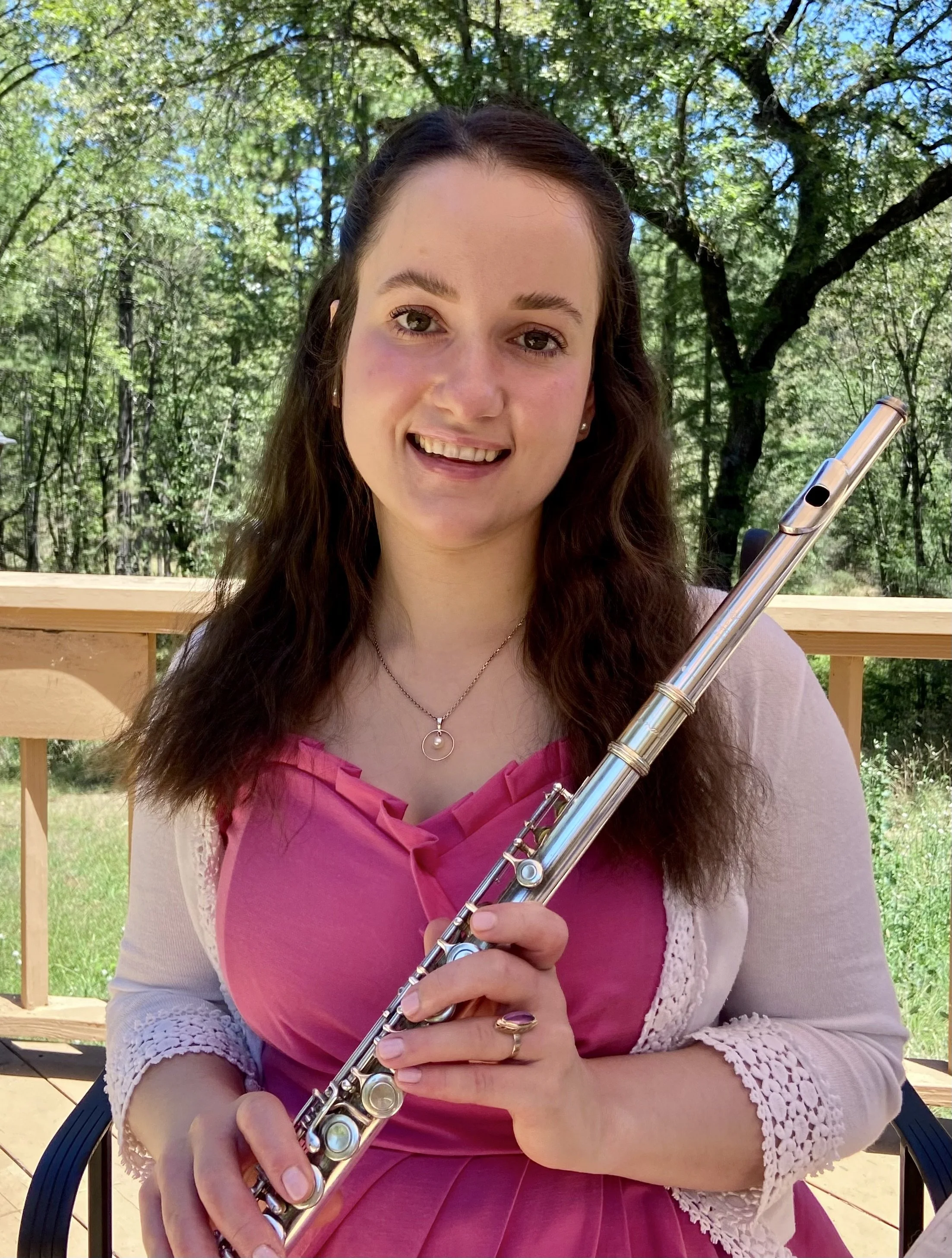 A young woman with long brown hair holding a flute, standing outdoors on a wooden deck with greenery and trees in the background, smiling at the camera.