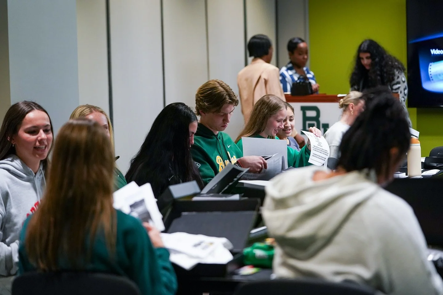 Group of students sitting at tables, engaging in activities, with some standing at the back in a classroom or meeting room.