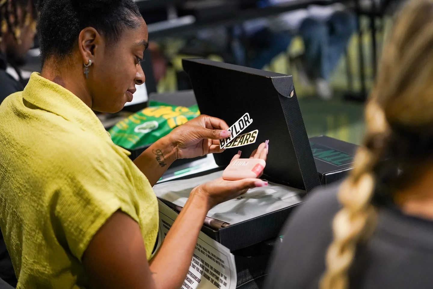 A woman in a yellow top opens a black box with stickers and pink items inside during a gathering, possibly an event or celebration.