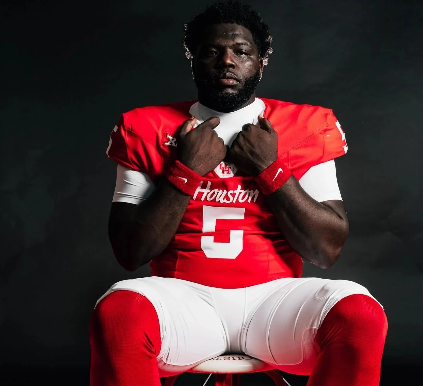 American football player in a red and white Houston jersey with the number 5, sitting against a dark background, crossing his arms over his chest.
