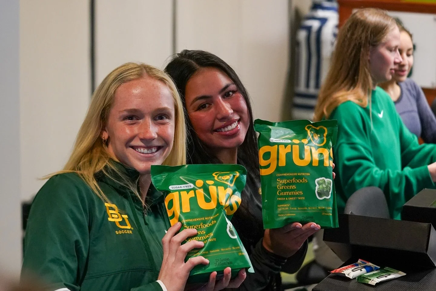 Two young women smiling and holding bags of Grün Superfoods Greens Gummies in an indoor setting, with other people in the background.