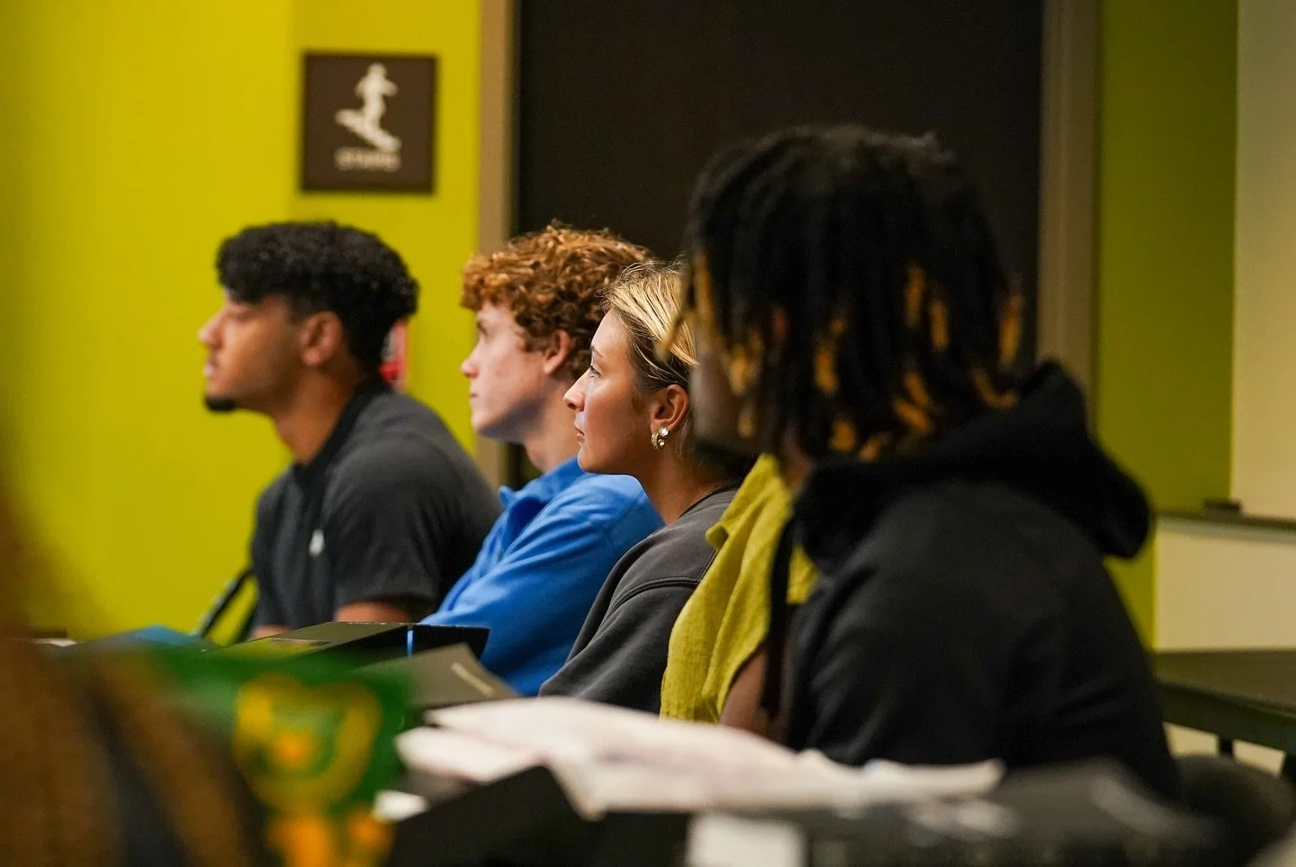 Four young adults sitting in a row, attentively listening in a classroom, with a green wall and framed artwork in the background.
