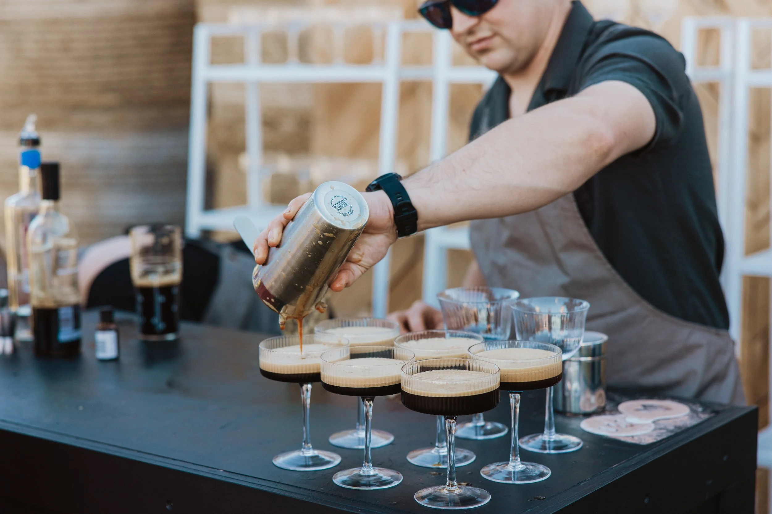 A man wearing sunglasses and a black shirt pours chocolate into glasses with layered dessert. There are several glasses with the dessert on a black table, along with bottles and bowls in the background.