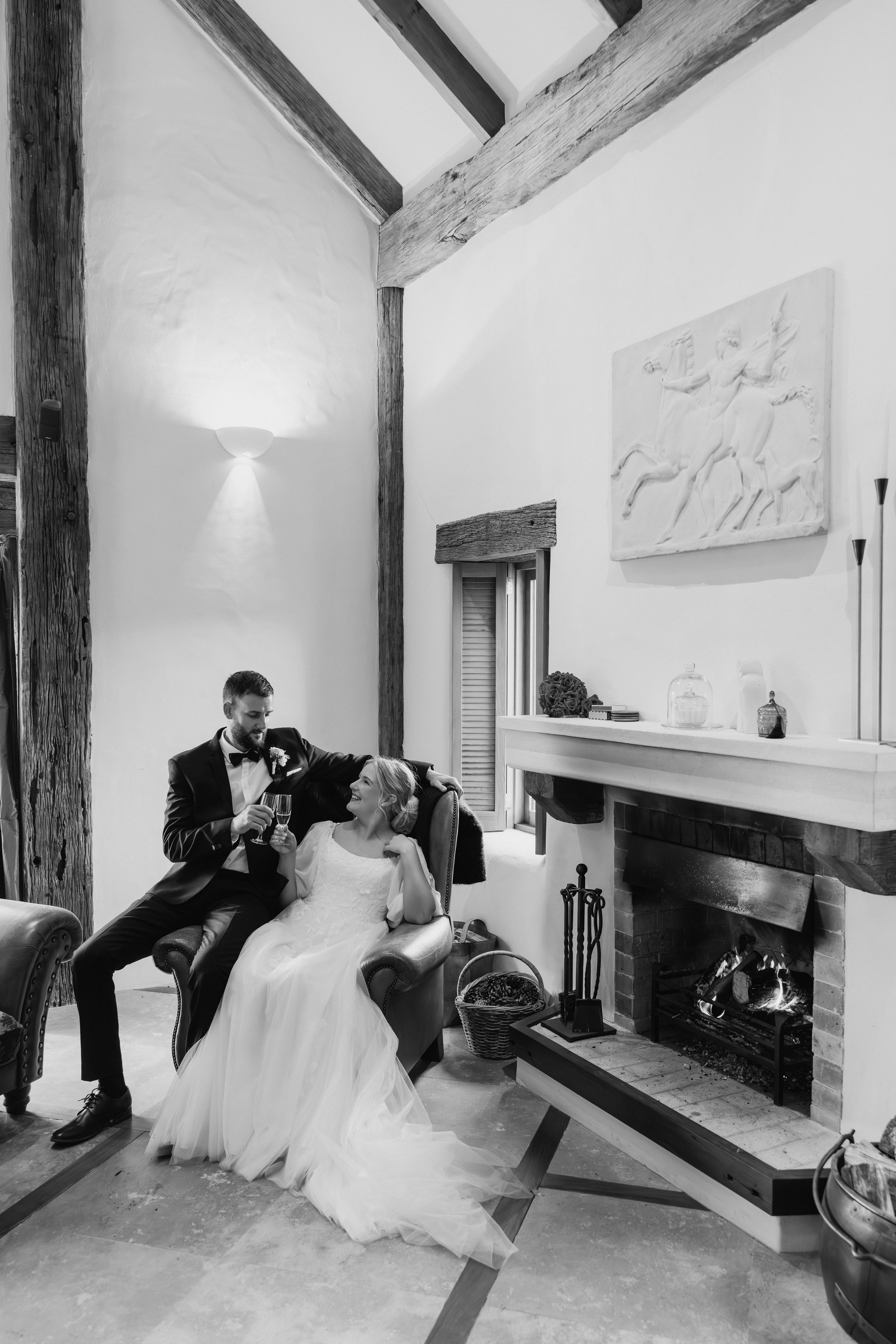 A bride and groom celebrating by a fireplace in a rustic room. The bride is seated in a chair wearing a white wedding dress, smiling, and looking up at the groom. The groom, dressed in a tuxedo with a bow tie, is standing beside her, holding a glass, while leaning slightly over her, smiling. The room has a large wooden beam structure, a painting of a horse on the wall, modern floor lamps, and a decorated mantel with storage jars and a bouquet of flowers.