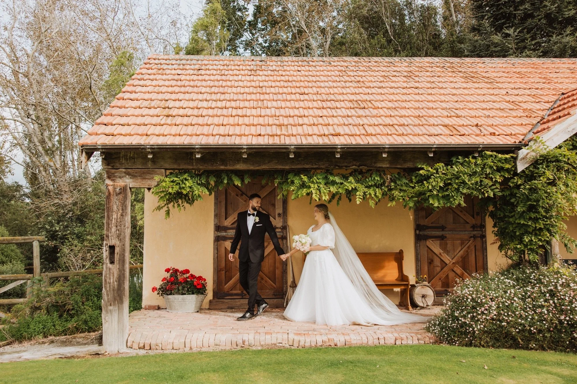 A bride and groom stand in front of a rustic building with a tiled roof, holding hands and smiling at each other during their wedding. The bride wears a white gown, and the groom is dressed in a black tuxedo. There are lush green plants and flowers around them.