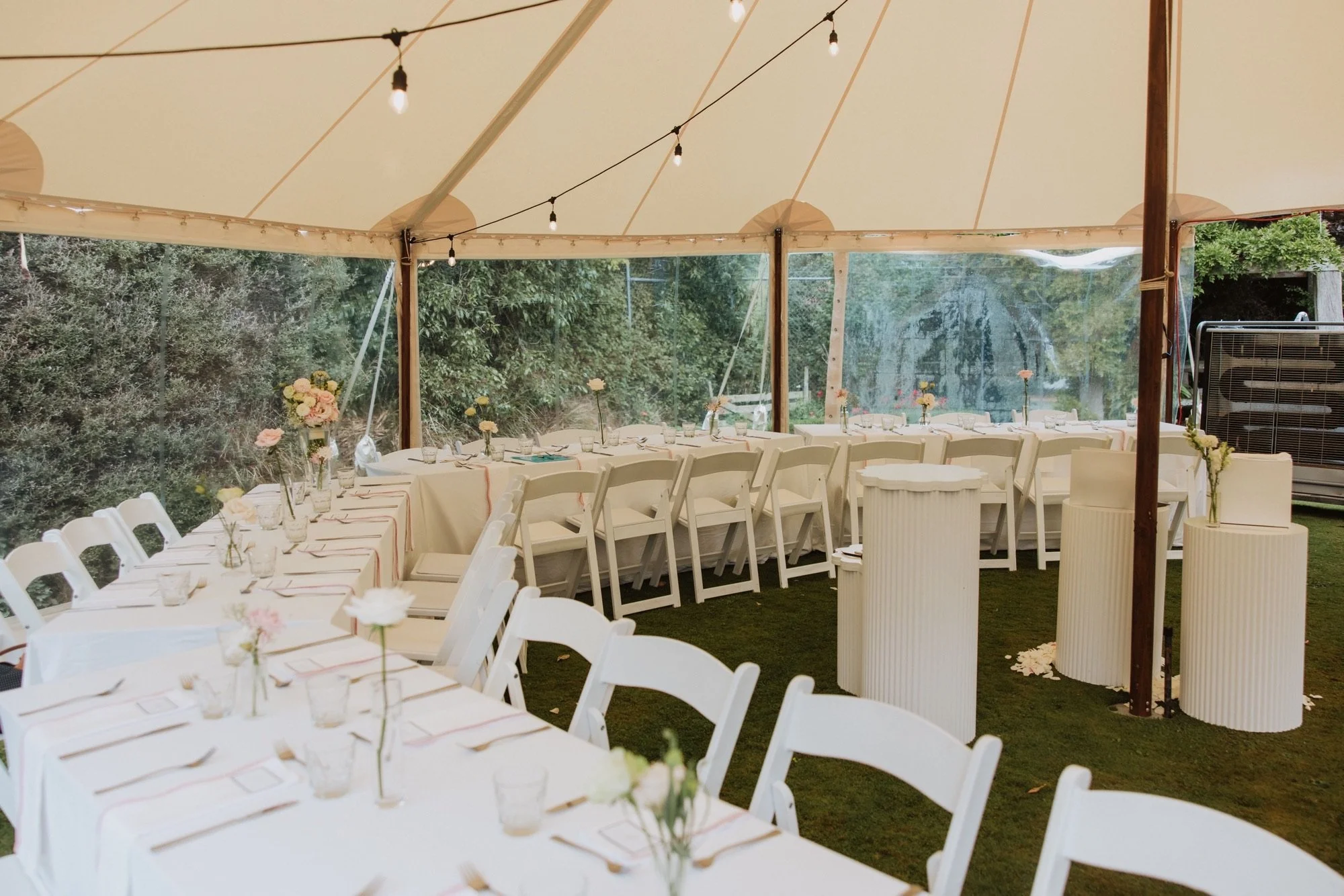 Outdoor event setup with long tables covered in beige tablecloths, decorated with small flower arrangements in glass vases, under a tent with string lights, surrounded by green trees and grass.