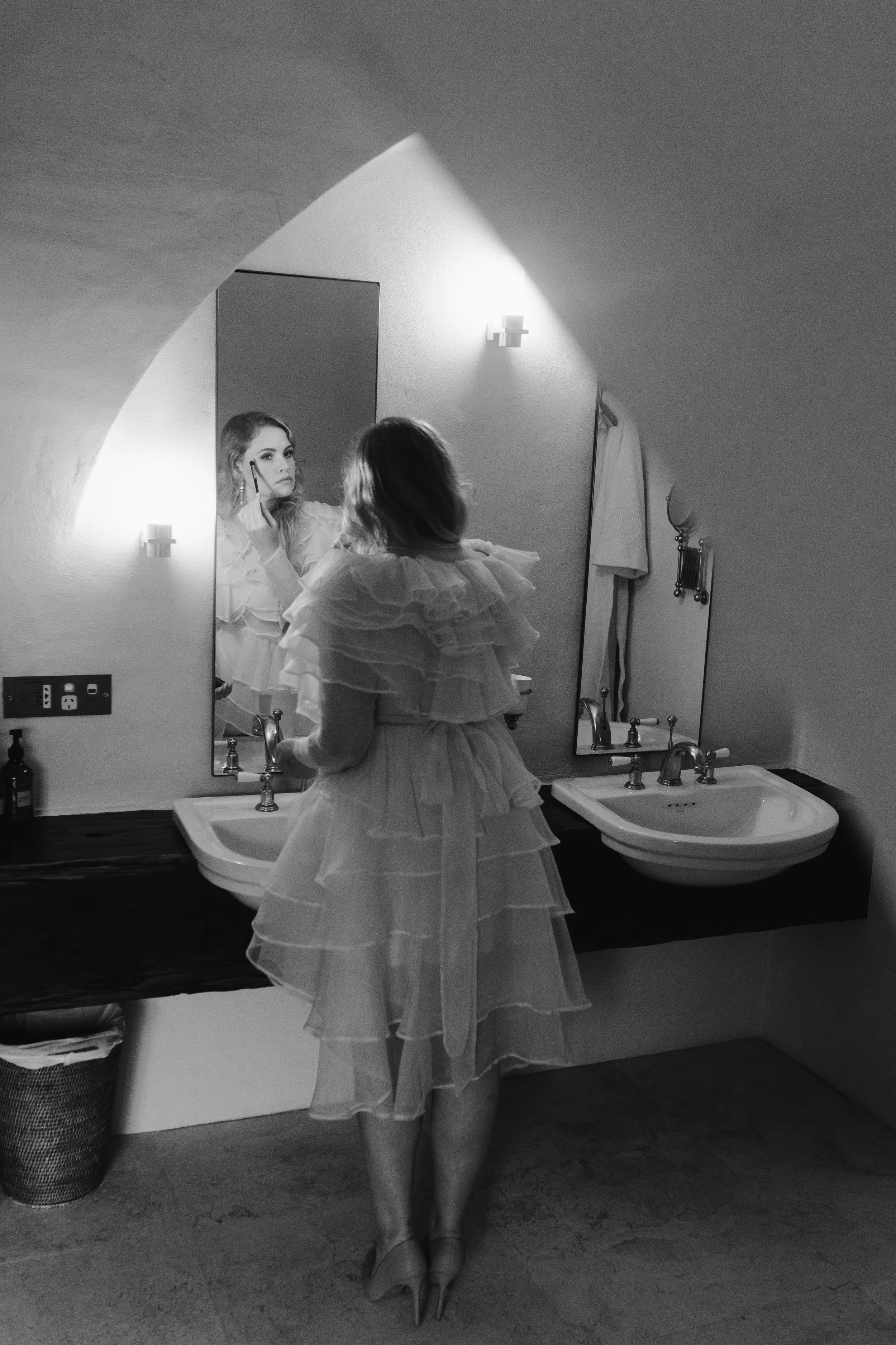 A woman in a layered, ruffled dress is standing in front of a mirror, applying makeup while looking at her reflection in a bathroom with dual sinks and a dark countertop.