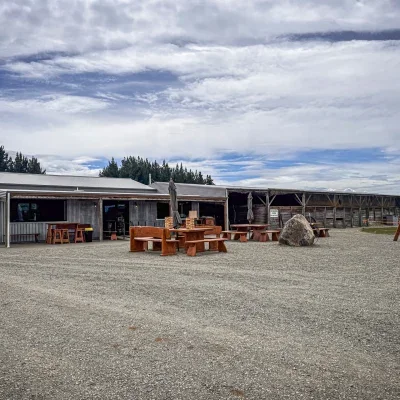 Outdoor seating area with wooden tables and benches on gravel, building with a covered patio, cloudy sky, trees in the background.