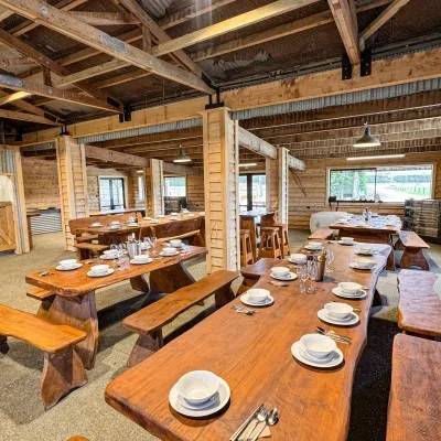 Interior of a rustic restaurant with wooden tables set for dining, featuring plates, glasses, and silverware, with exposed wooden beams on the ceiling and large windows.