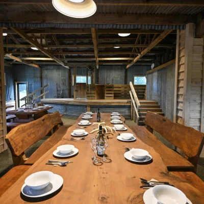 A rustic dining area with a long wooden table set with white plates, bowls, glasses, and cutlery, inside a barn-like structure with wooden beams and metal siding.
