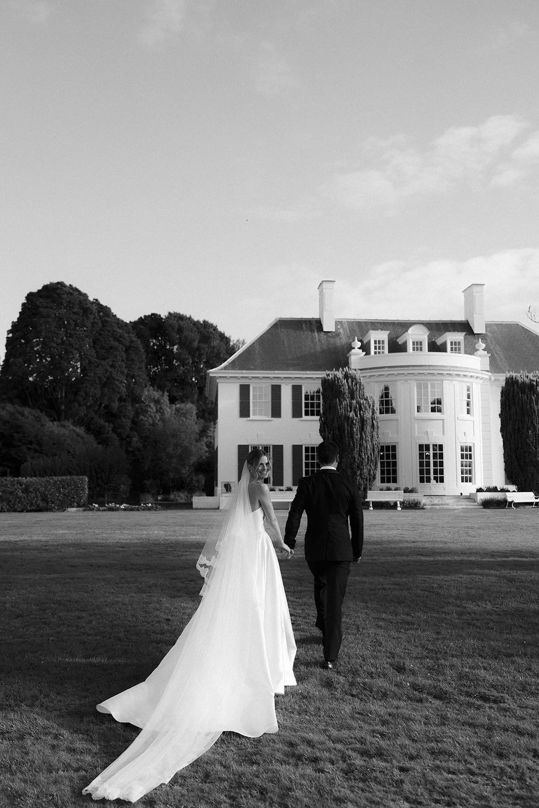 A bride and groom walk hand in hand across a grassy lawn in front of a large, white mansion during a wedding photoshoot, black and white photograph.