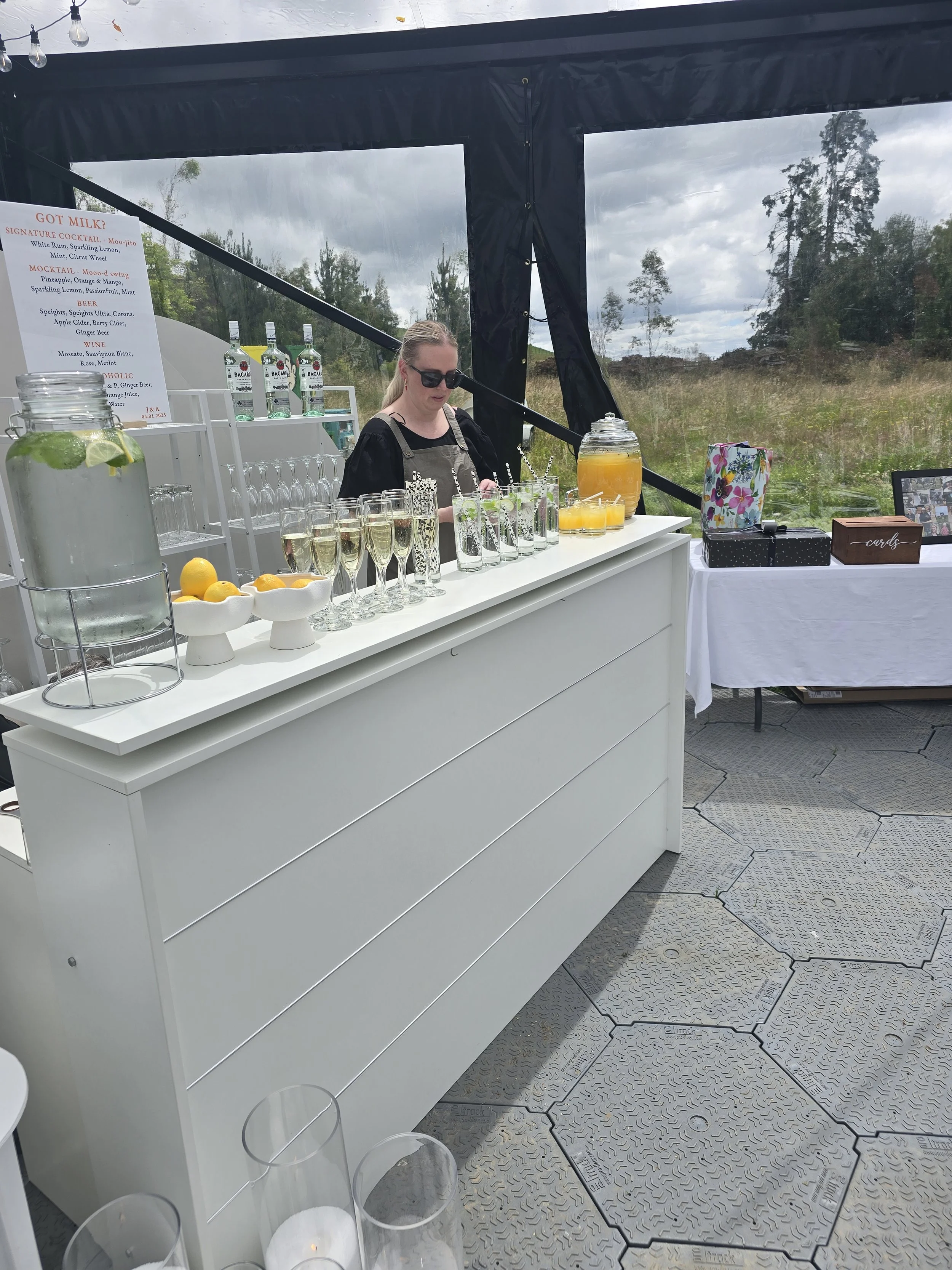 A bartender preparing drinks at an outdoor bar with a scenic view of trees and cloudy sky in the background. There are bottles of Bacardi rum, glasses of champagne, lemons, and a jar of orange juice on the bar. A sign with drink options and a box of greeting cards is visible on a table to the right.