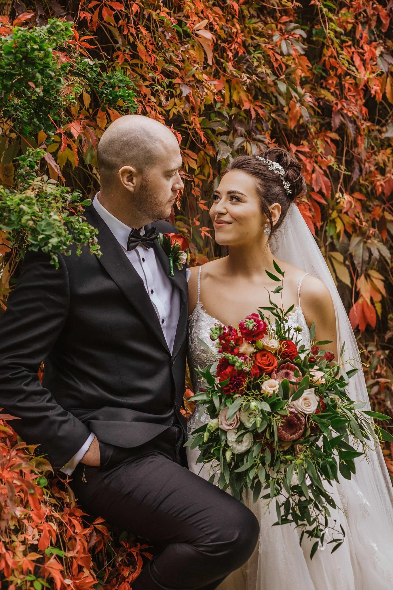 A bride and groom on their wedding day surrounded by orange and red fall foliage. The bride holds a large bouquet of red, pink, and white flowers with greenery, and wears a white lace wedding dress and a sparkling headpiece. The groom wears a black tuxedo with a bow tie and a boutonniere matching the bride's bouquet.