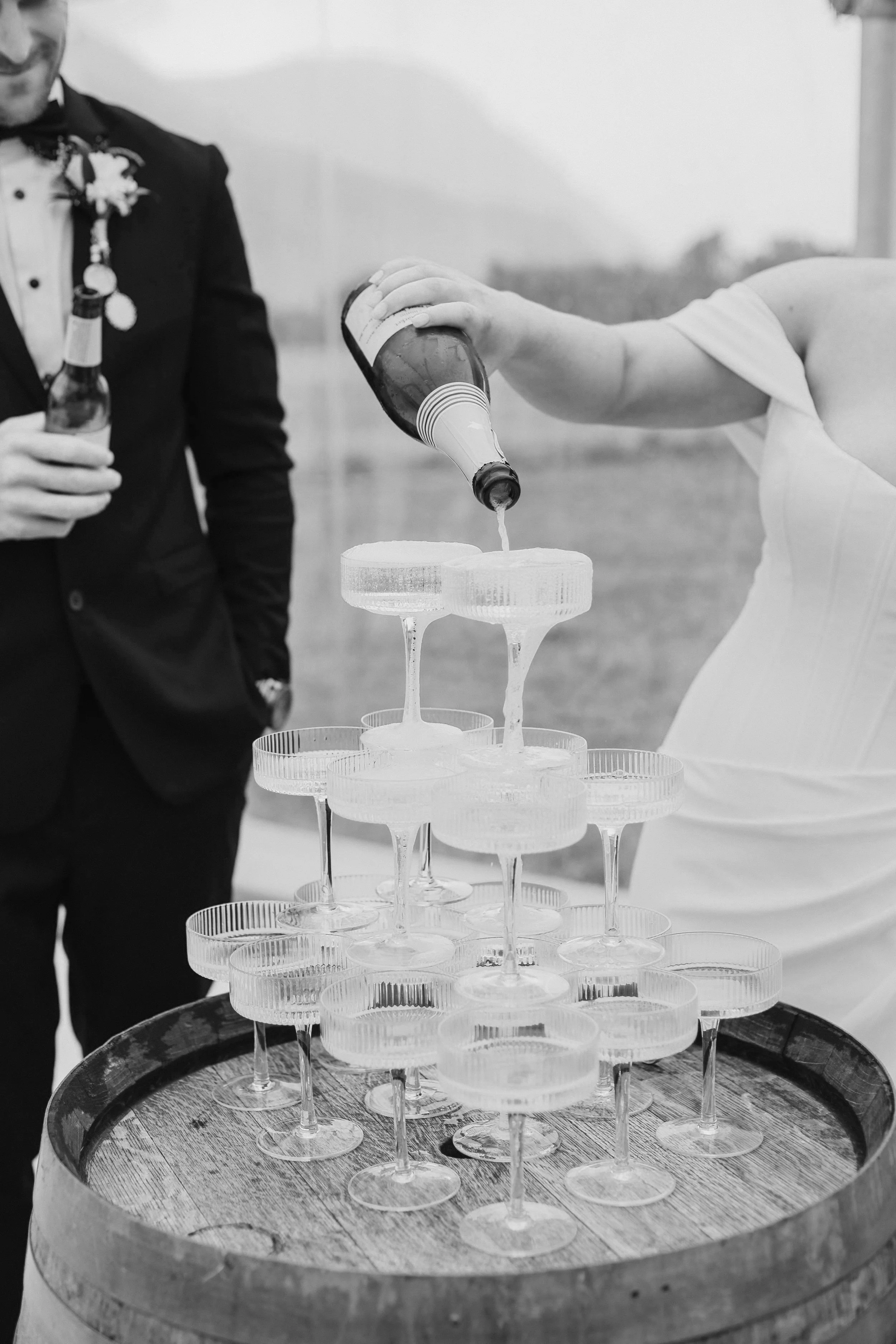 A woman in a wedding dress pours a drink into a champagne tower at a wedding reception, with a man dressed in a tuxedo holding a beer in the background.