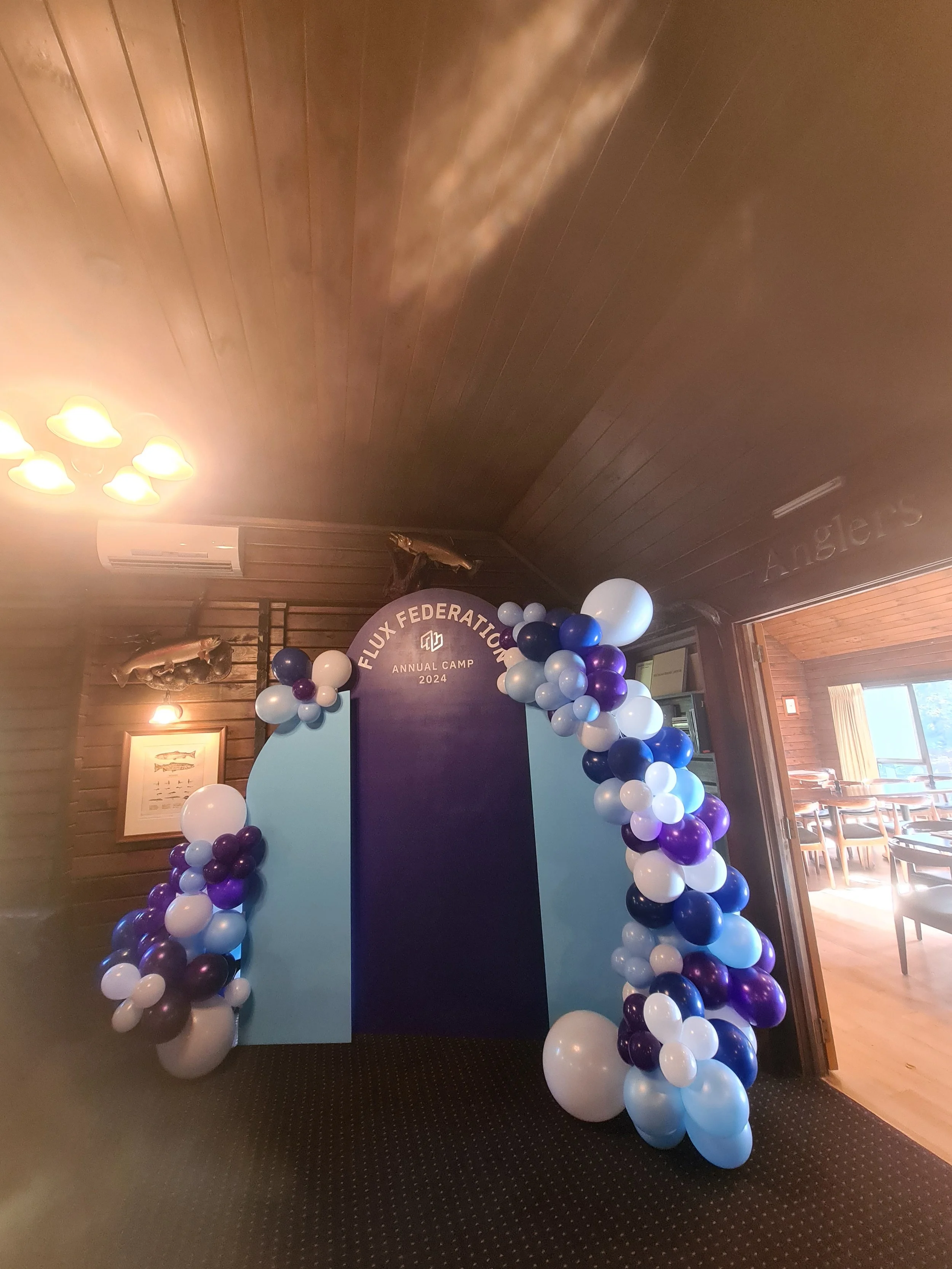 Balloon arch in purple, white, and blue colors framing a sign for Flux Federation's annual camp 2024, inside a room with wooden walls and a view of a dining area.