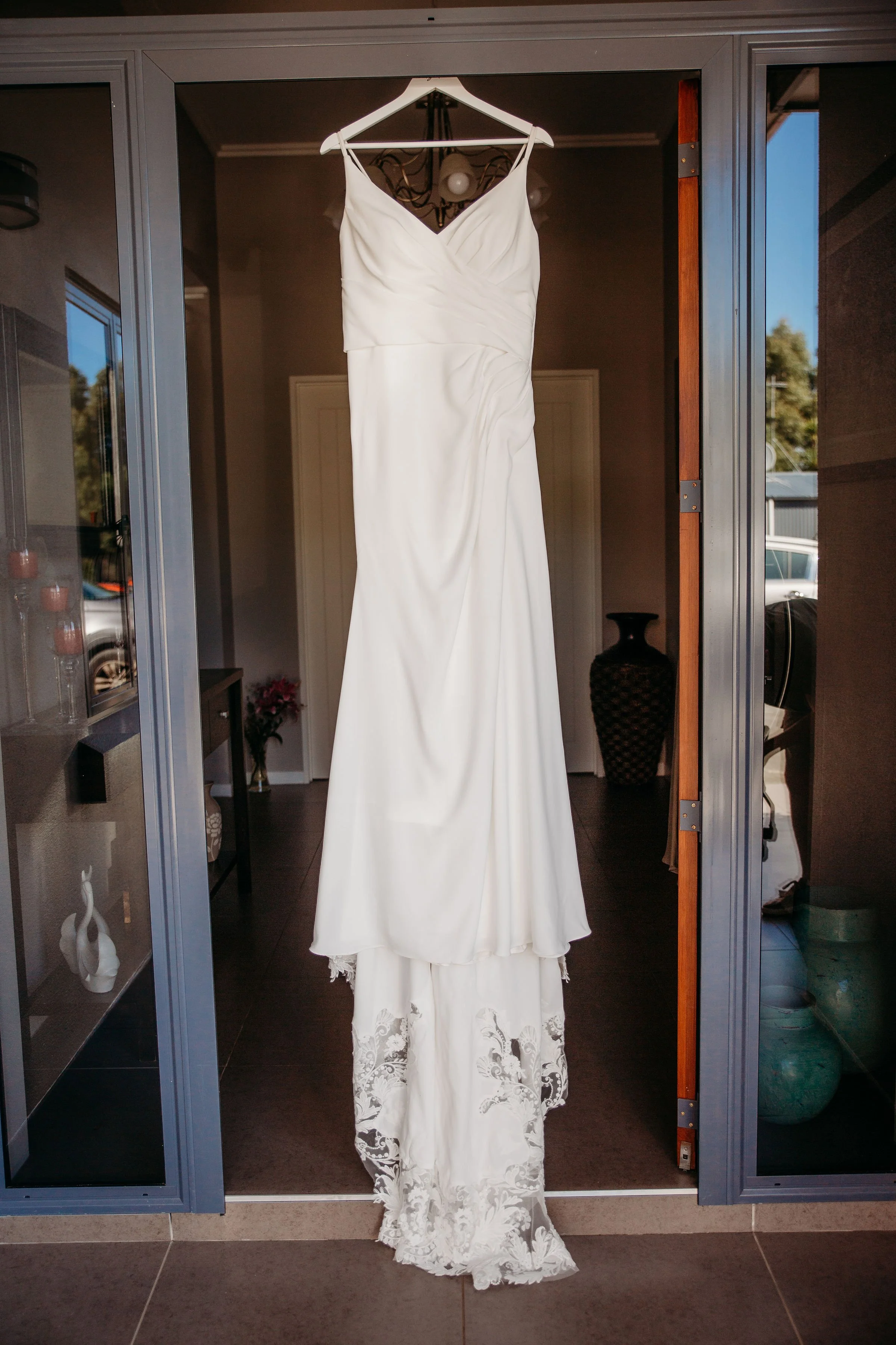 A white wedding dress with lace detailing at the bottom hangs on a hanger inside a glass closet. The dress is displayed in front of a background with decorative vases, a floral arrangement, and a dark-colored vase. The closet doors are open, revealing the interior and the dress.