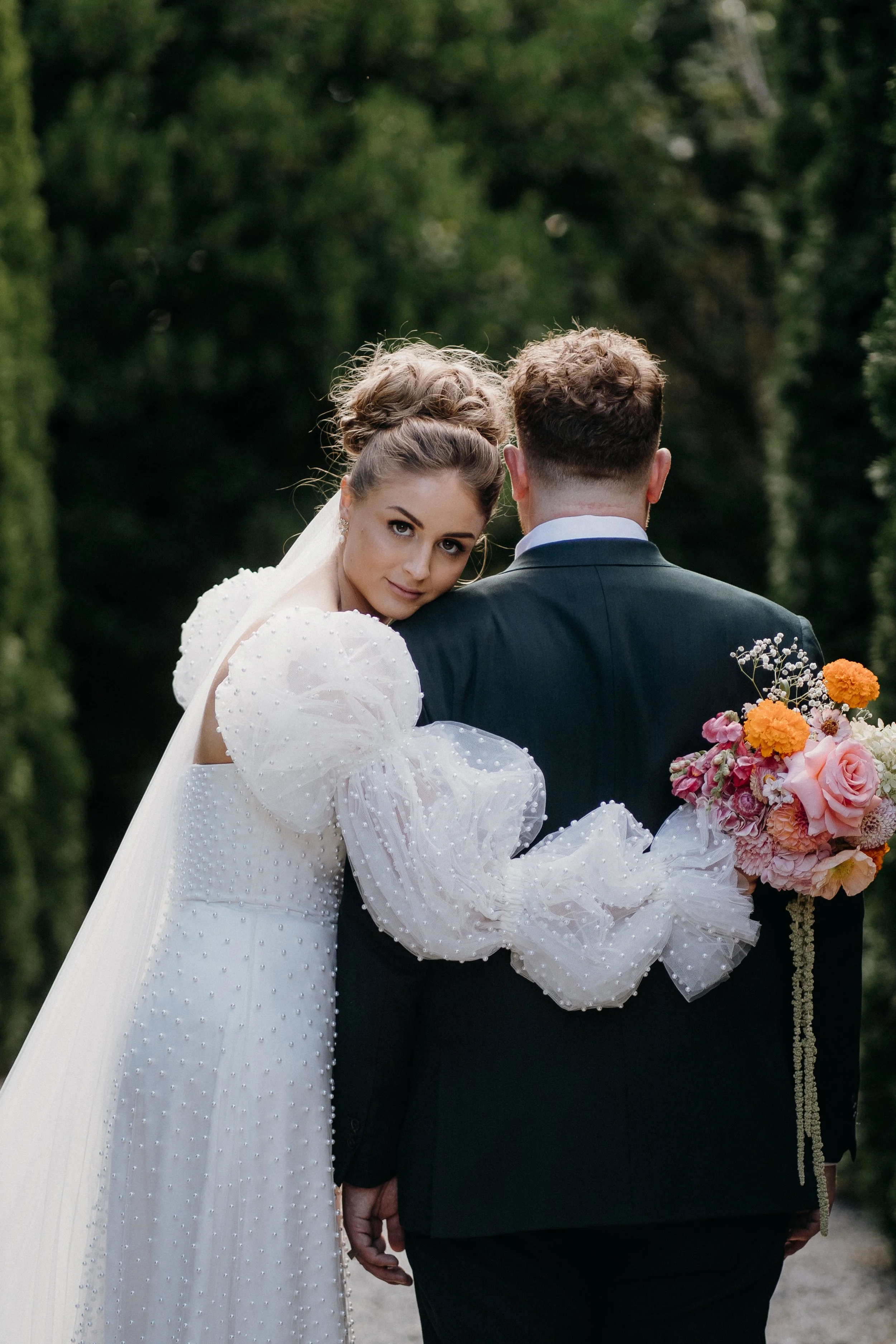 A bride and groom walking together outdoors, with the bride holding a bouquet of pink and orange flowers, the bride wearing a white dress with pearl details, and the groom dressed in a dark suit.