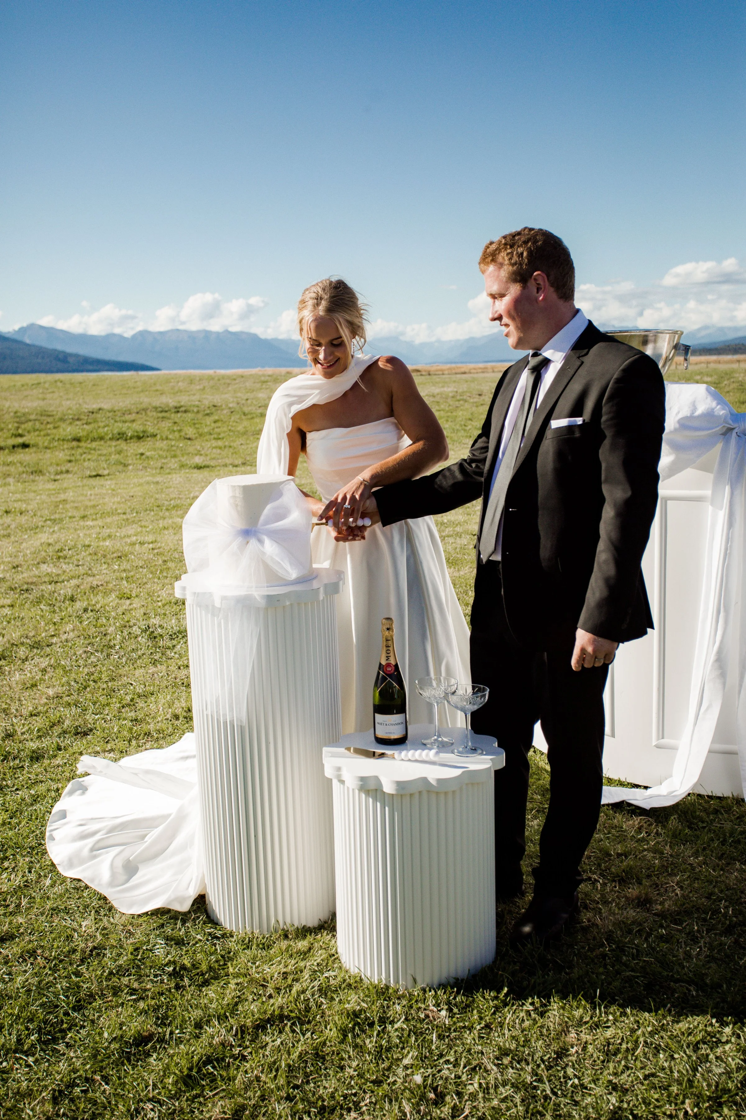 A bride and groom have a wedding ceremony outdoors on a grassy field with mountains in the background. The bride is smiling and wearing a white wedding dress, while the groom is dressed in a black suit. They are standing in front of a small table with a champagne bottle and glasses, and a white cloth and ribbons decorate the scene.