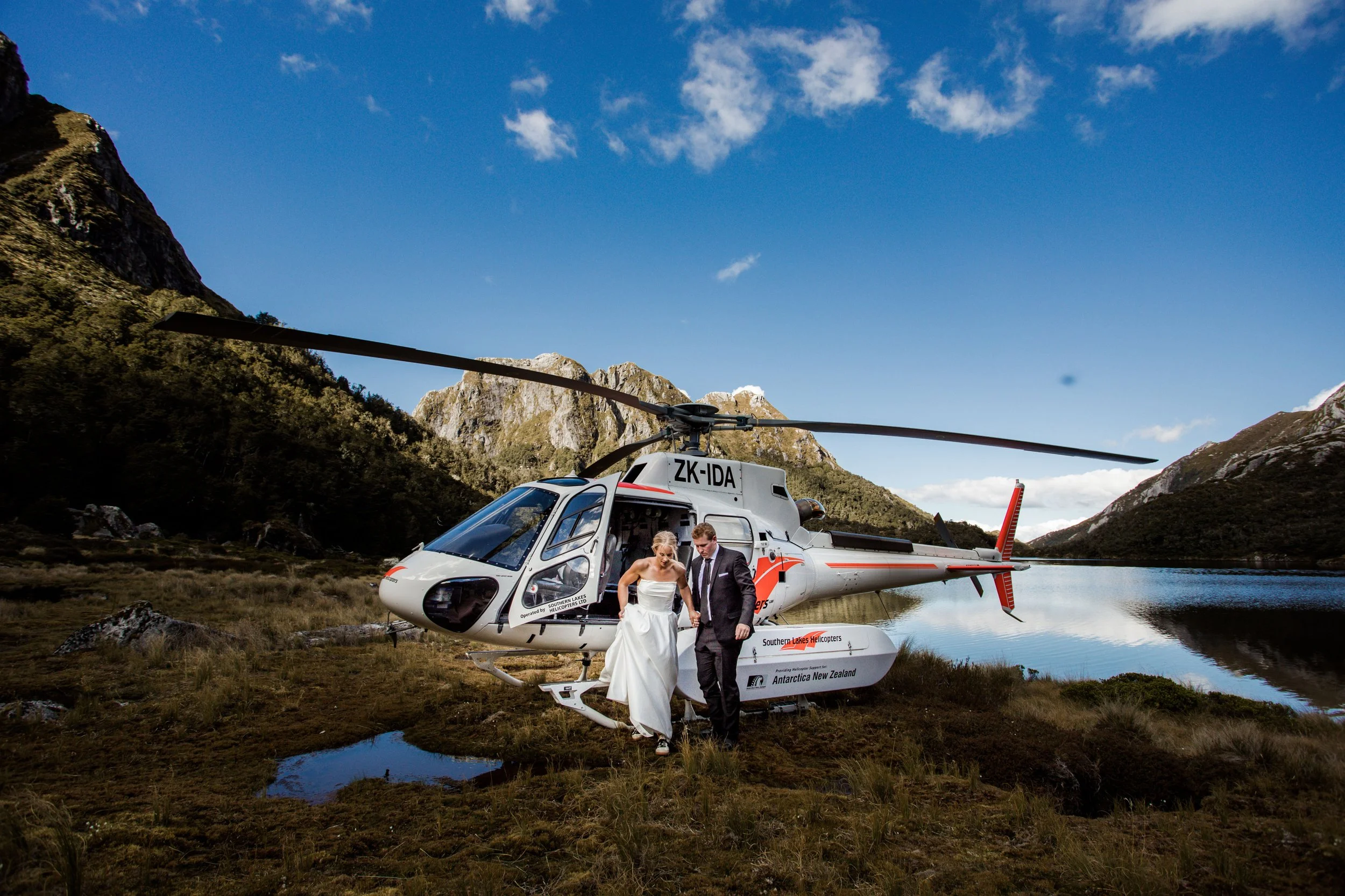 A helicopter with the registration ZK-IDB landed near a mountain lake, with a bride and groom in wedding attire walking away from it.