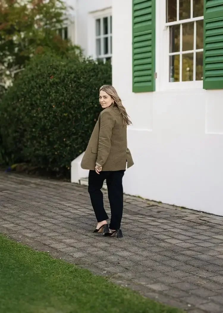 A woman is standing on a paved walkway next to a white house with green shutters, wearing a brown blazer, black pants, and high-heeled shoes, looking back at the camera with a smile.