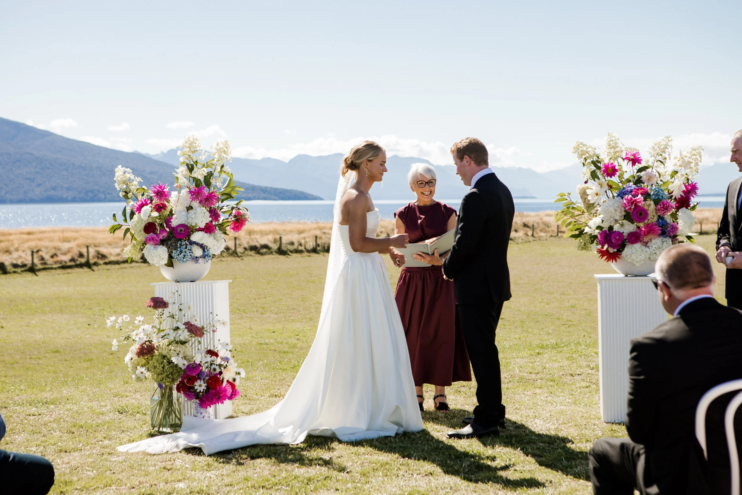 A couple getting married outdoors by a lake with mountains in the background. The bride is in a white gown and the groom in a black suit. An officiant stands between them holding a book. Two large floral arrangements are on white pedestals on either side of the couple.