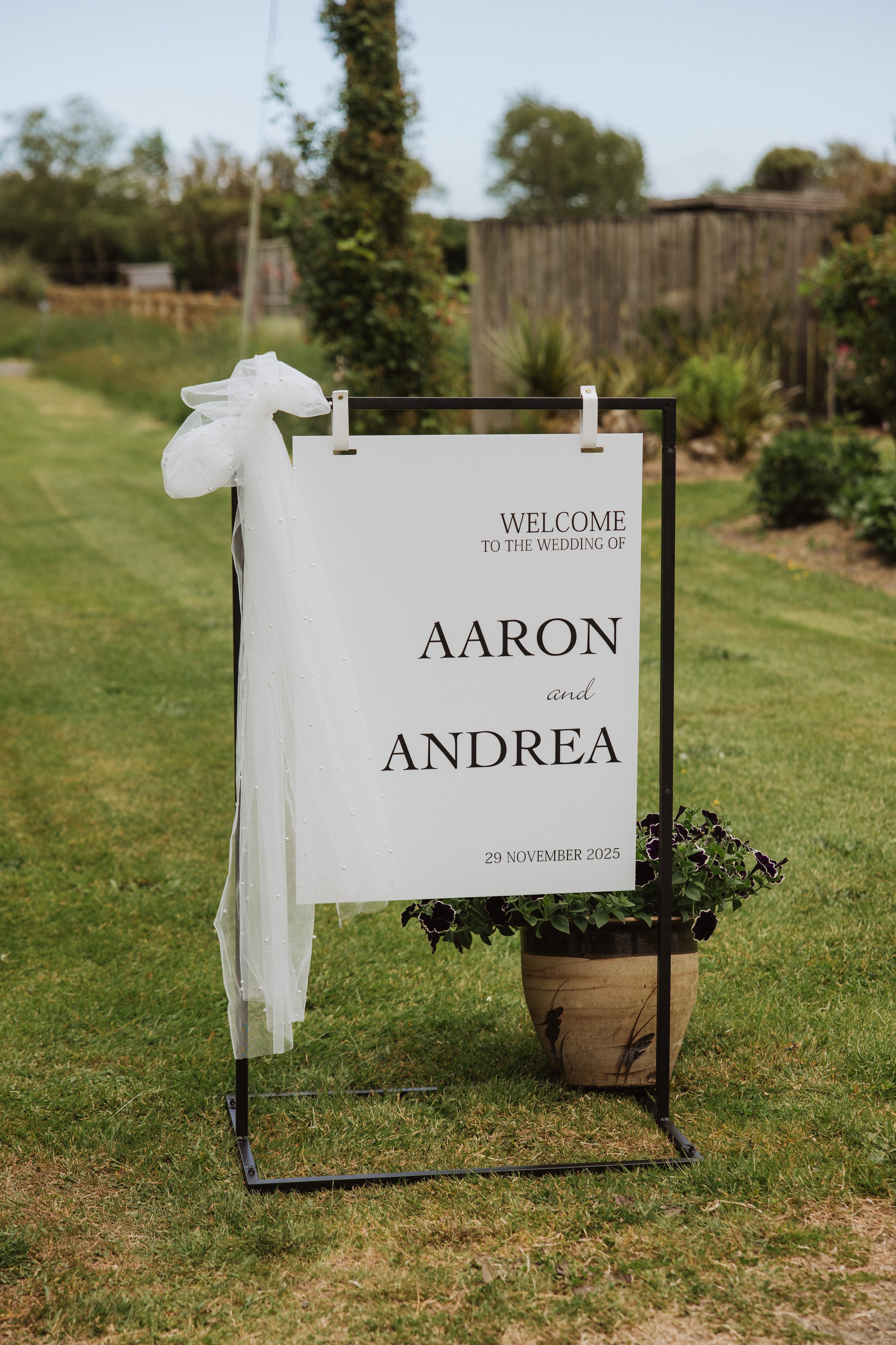 A welcome sign at a wedding with the names Aaron and Andrea, dated November 29, 2025, outdoors on a grassy area, decorated with a white ribbon.