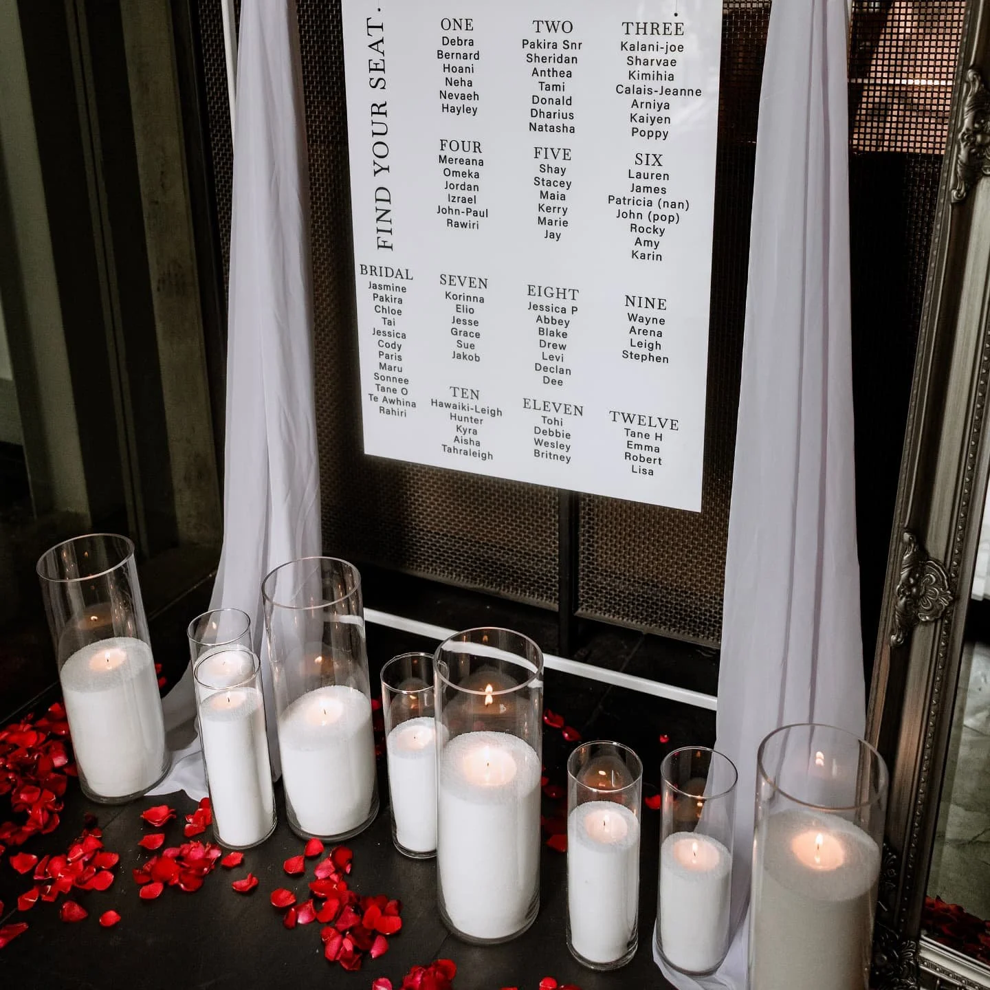 A wedding seating chart displayed on a stand draped with white fabric, surrounded by candles and scattered red rose petals, with a mirror frame on the right side.