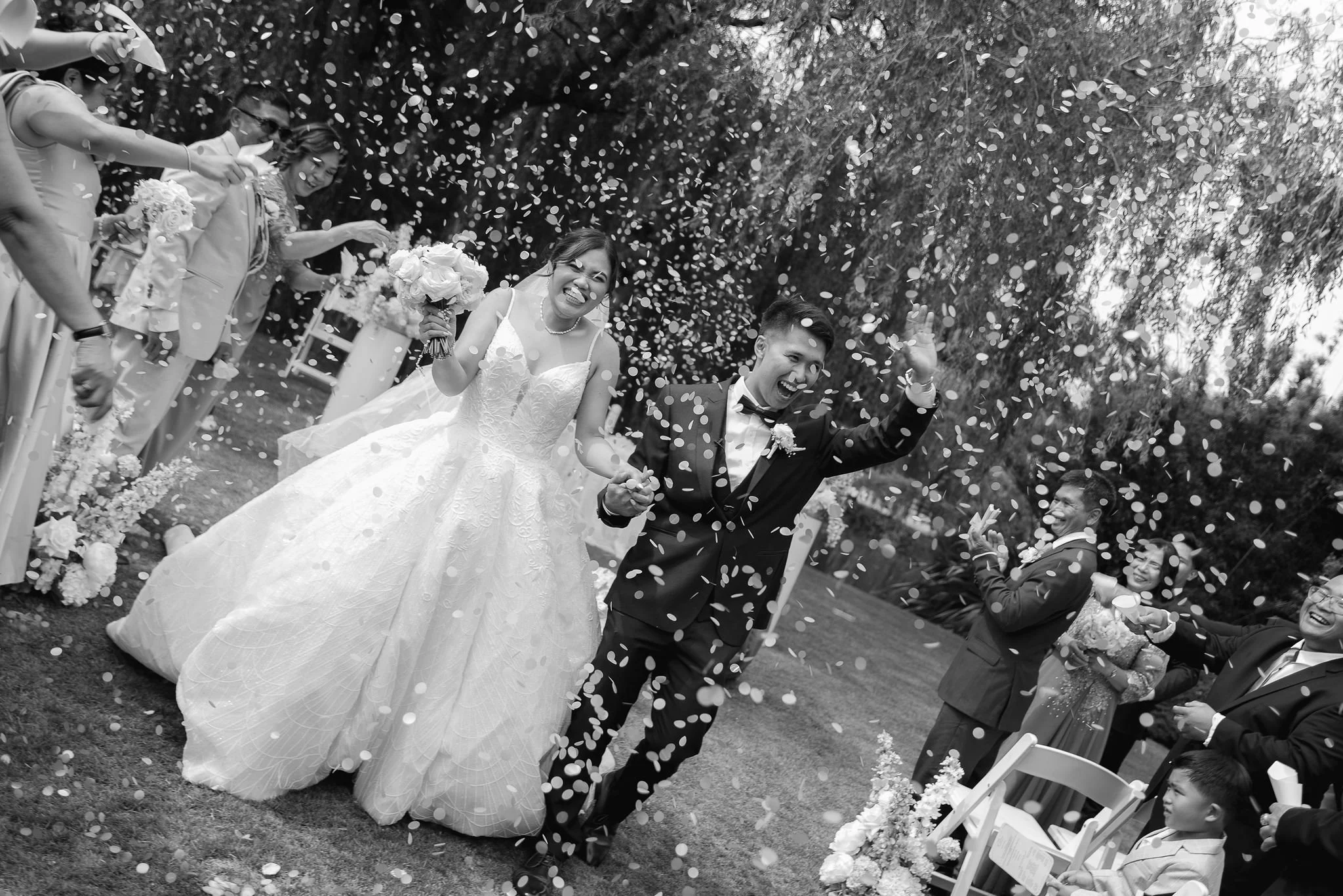 A bride and groom celebrating outdoors at a wedding, surrounded by guests throwing confetti.