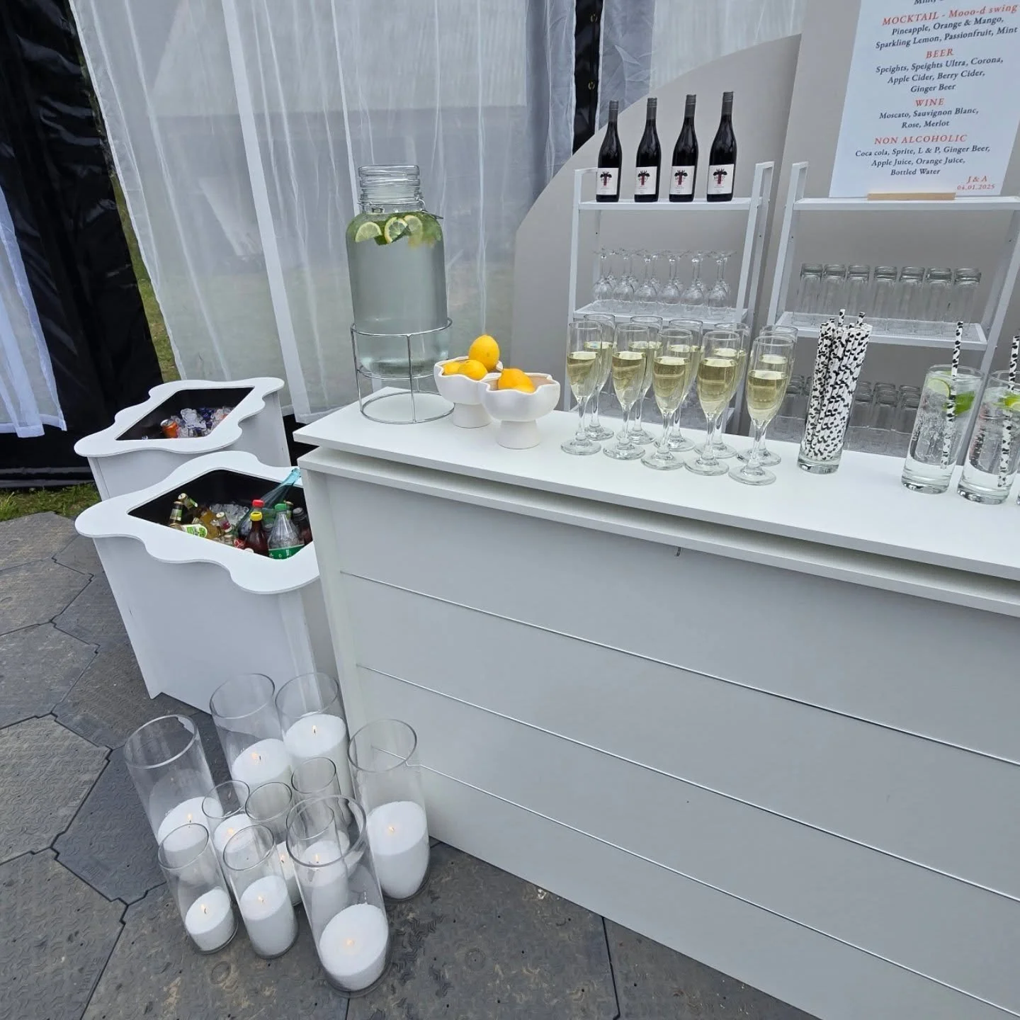 White bar with alcoholic and non-alcoholic beverages, including wine and sparkling water, set up outdoors with glasses and candles, and a menu board in the background.
